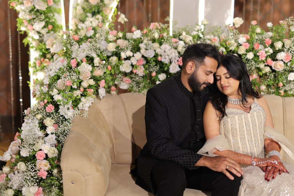 A couple in elegant attire sits intimately on a sofa surrounded by lush pink and white floral arrangements.