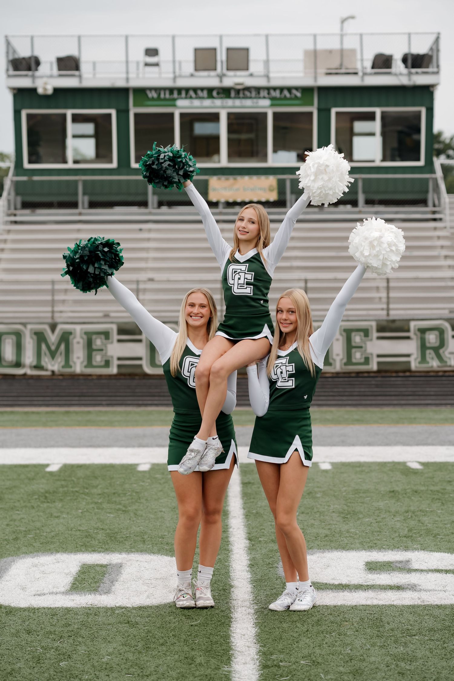 Three cheerleaders in green uniforms perform a stunt on a football field, with one lifted overhead holding white pom-poms.