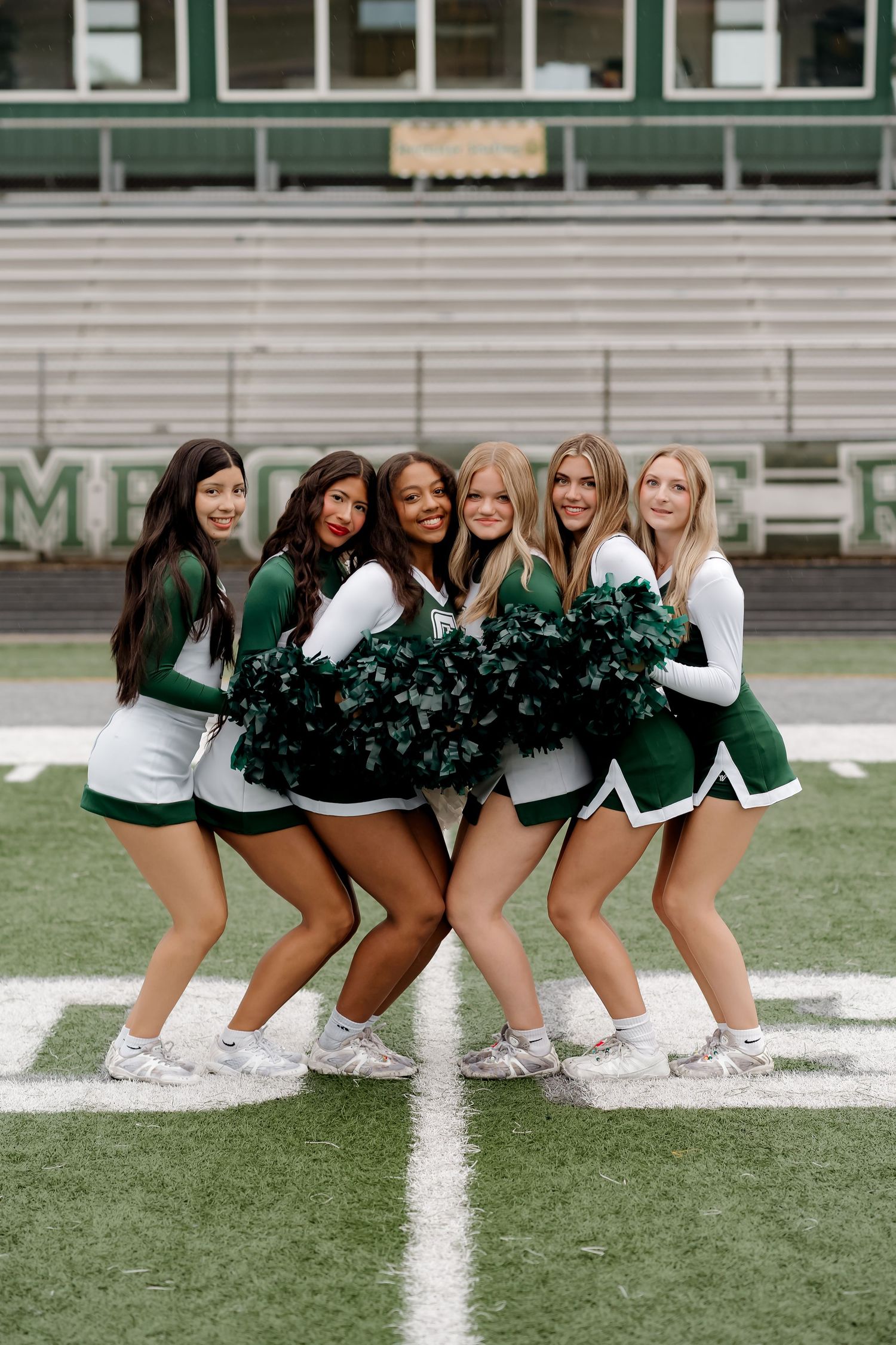 Six cheerleaders in green and white uniforms pose with pom-poms on a football field stadium.