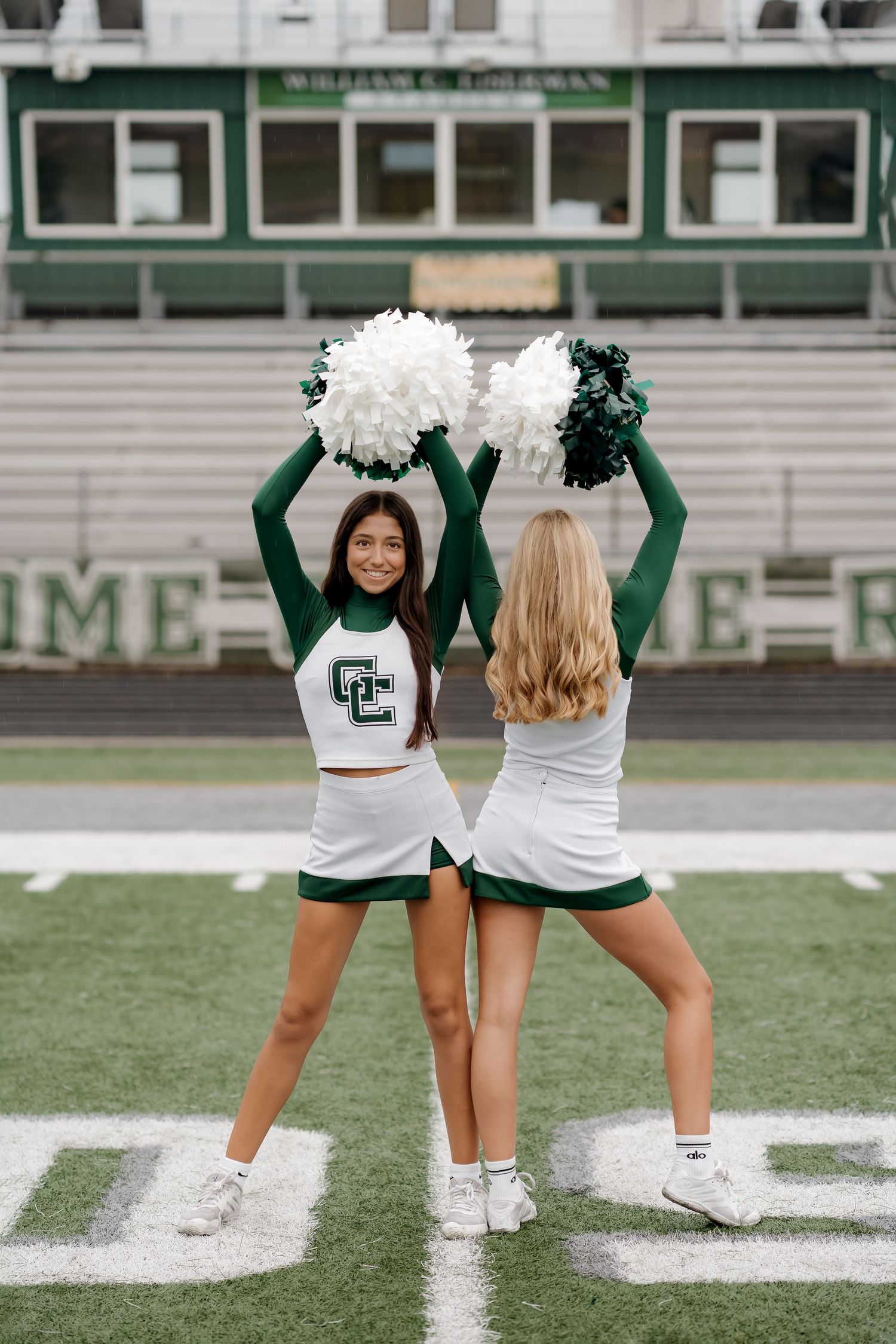 Two cheerleaders in green and white uniforms hold pom-poms overhead on a football field.