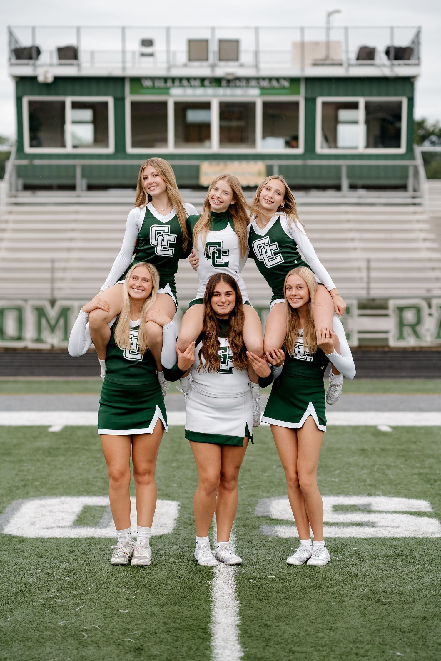 Cheerleaders in green and white uniforms pose in a pyramid formation on a football field.