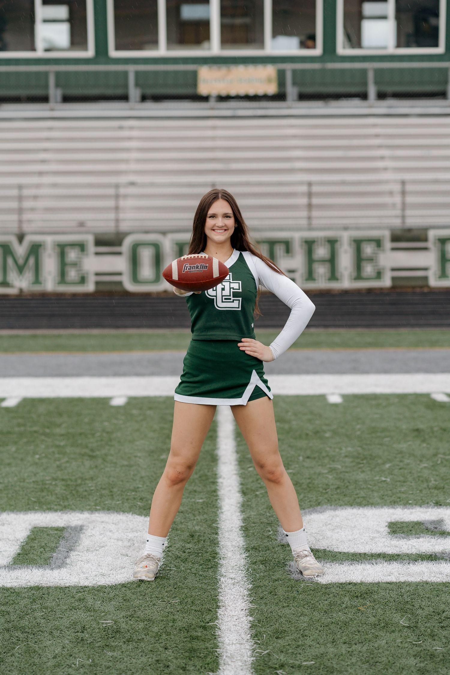 Cheerleader in green uniform holds football on field.