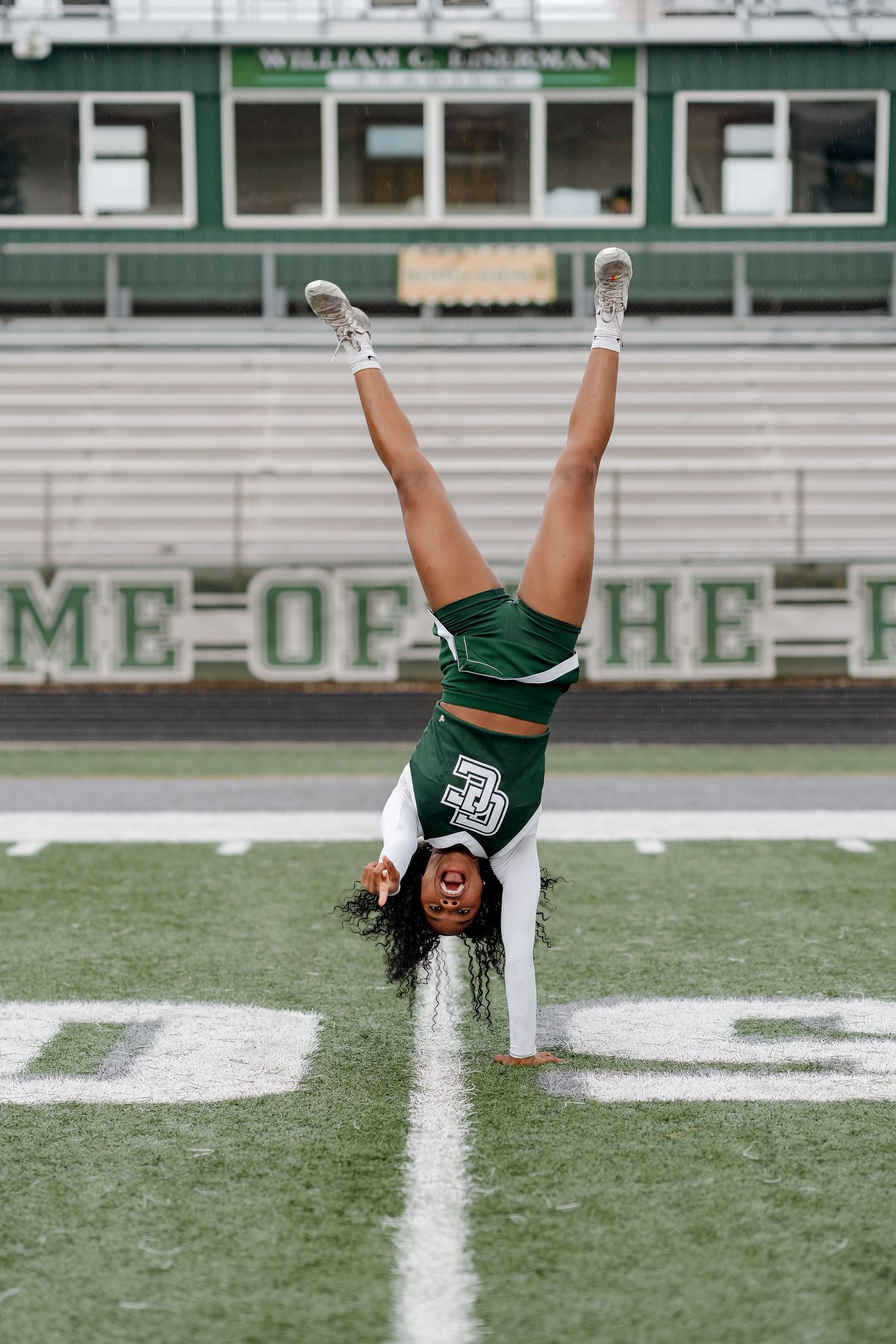 Cheerleader performs a handstand on a green football field wearing a green and white uniform with legs raised high.
