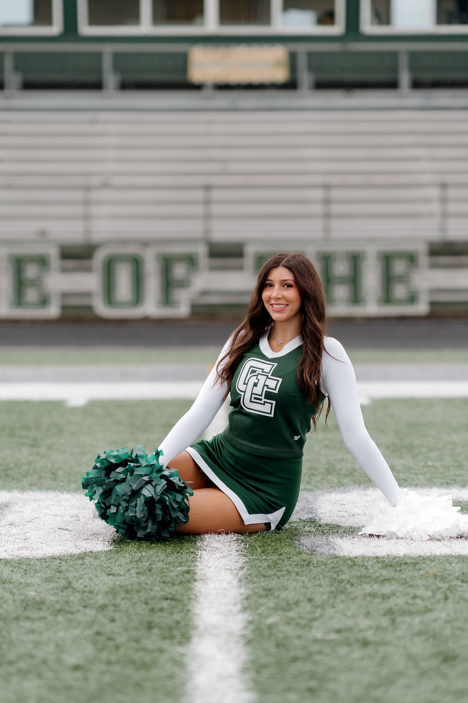 A cheerleader in a green uniform sits on a football field holding green pom-poms, smiling confidently.