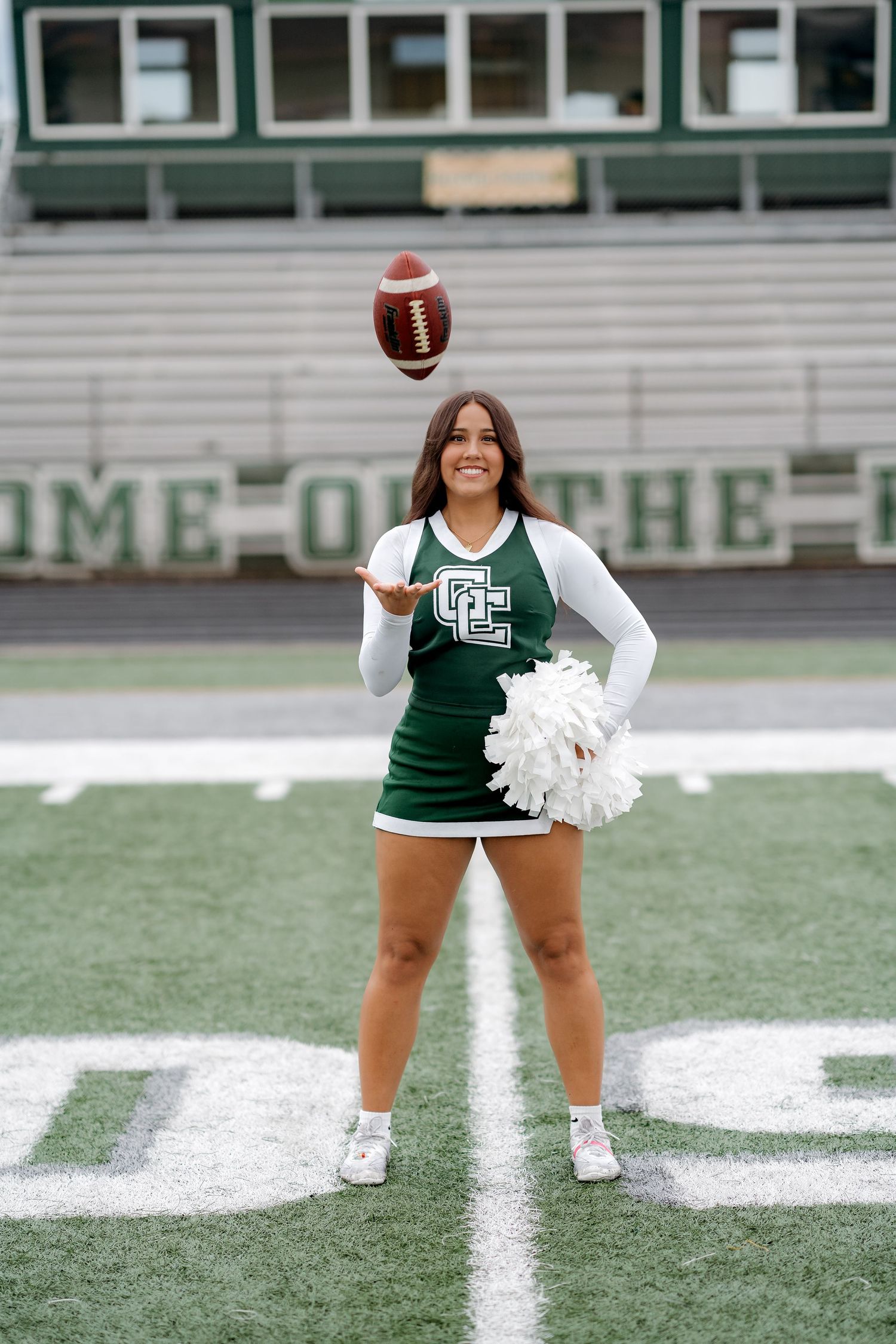 Cheerleader in green uniform tossing football on field, holding white pom-poms, stadium bleachers in background.