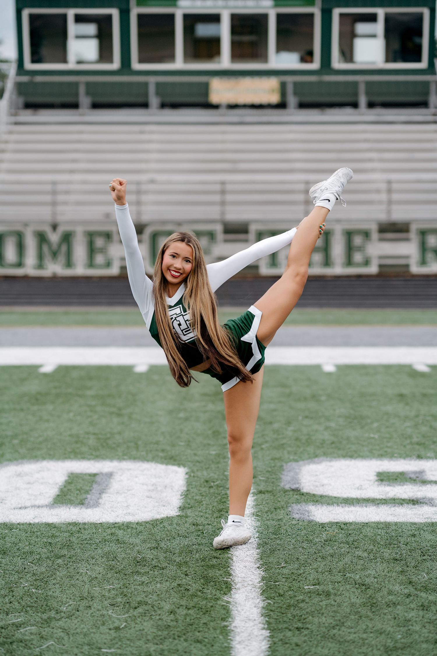 A GCHS Rams cheerleader performs a high kick on the football field in a green and white uniform.