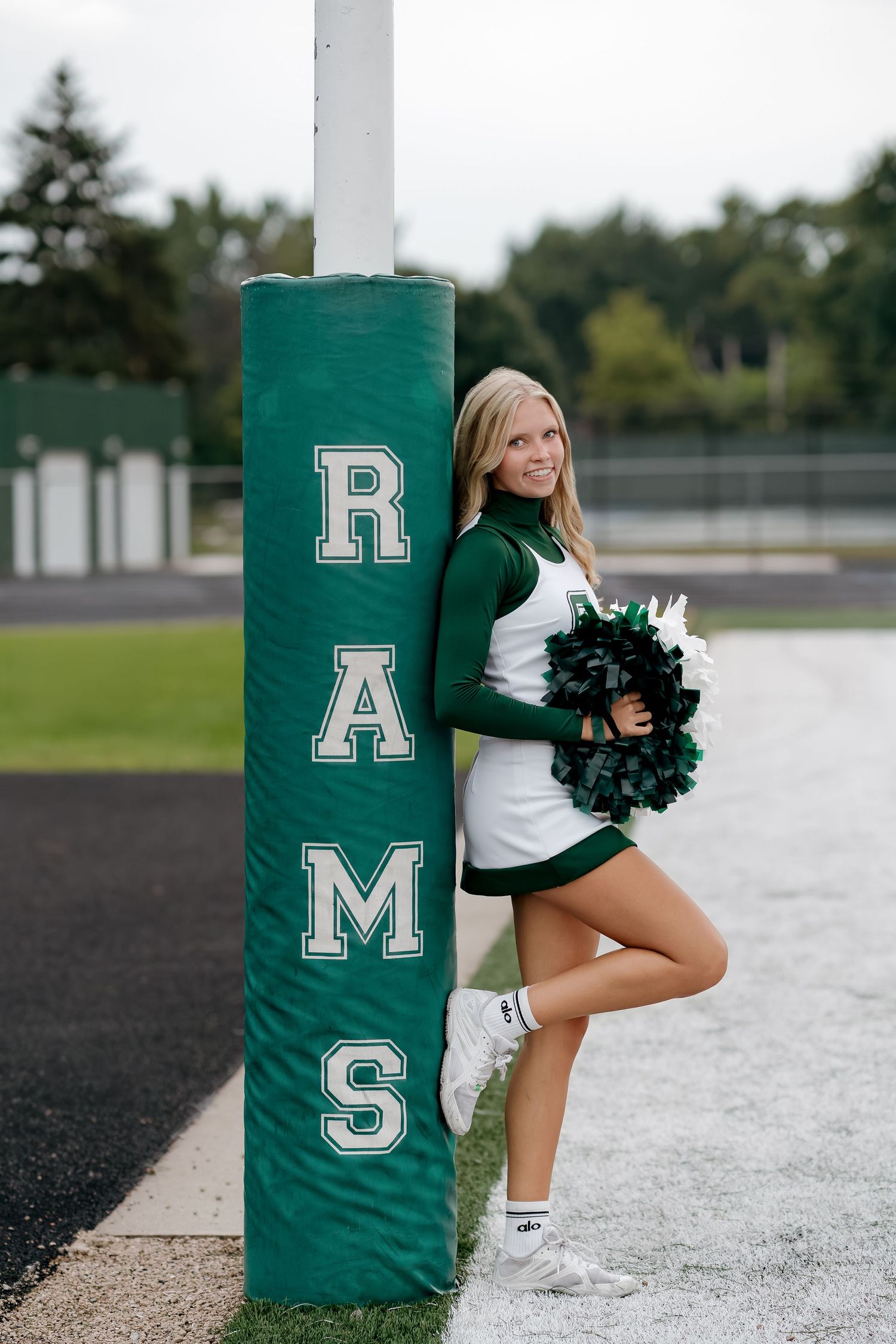 Blonde cheerleader in green and white uniform holding pom-poms beside a Rams goalpost pad on a football field.
