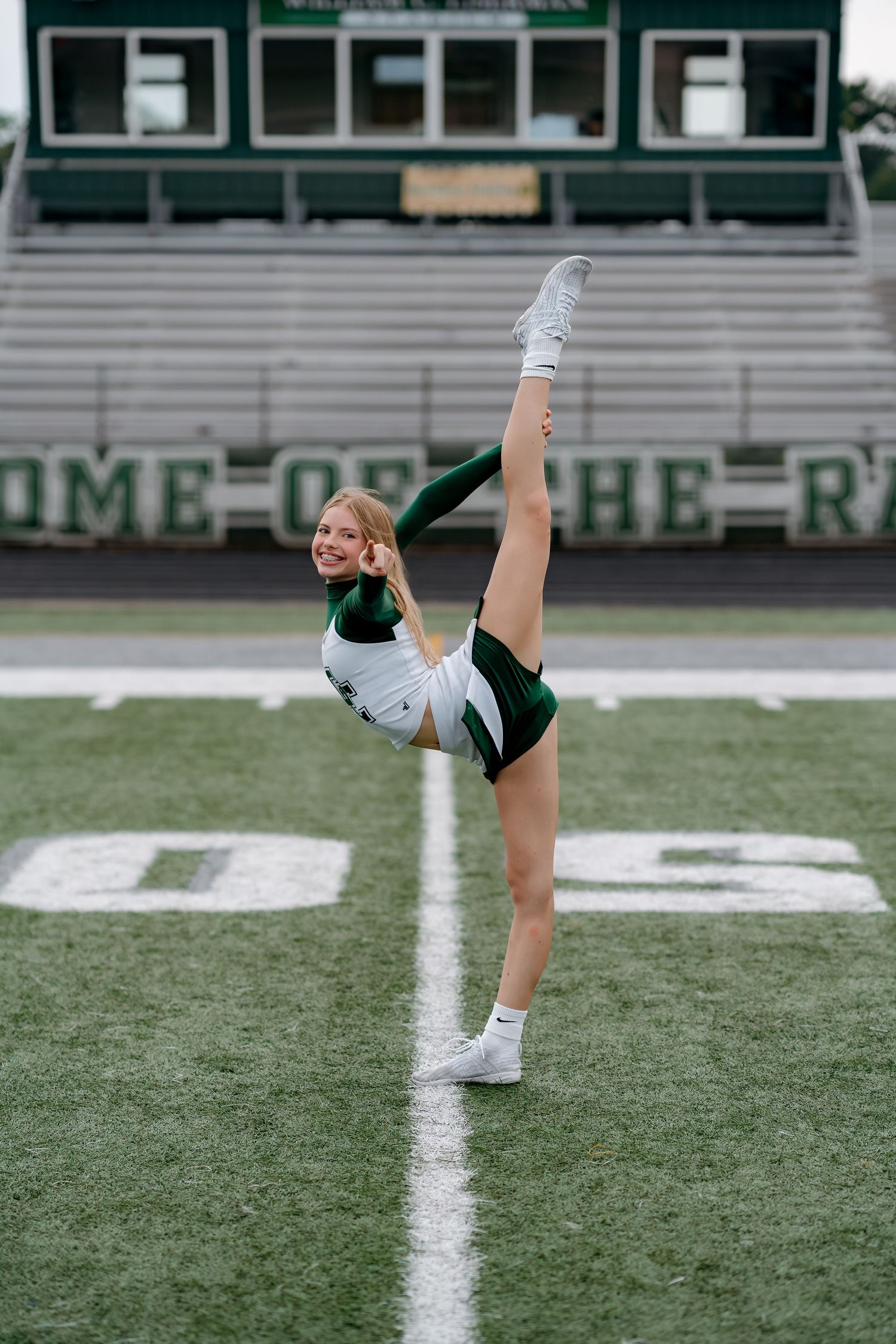 A cheerleader in a green uniform performs a high kick on a football field with stadium bleachers in the background.