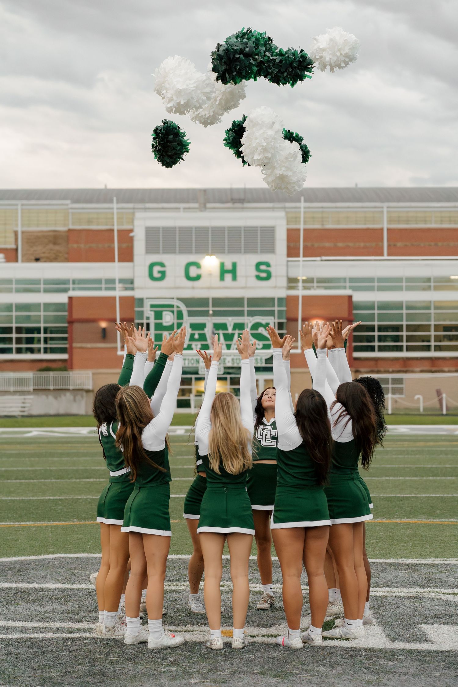 GCHS Rams cheerleaders in green uniforms tossing white and green pom-poms in the air on the football field.