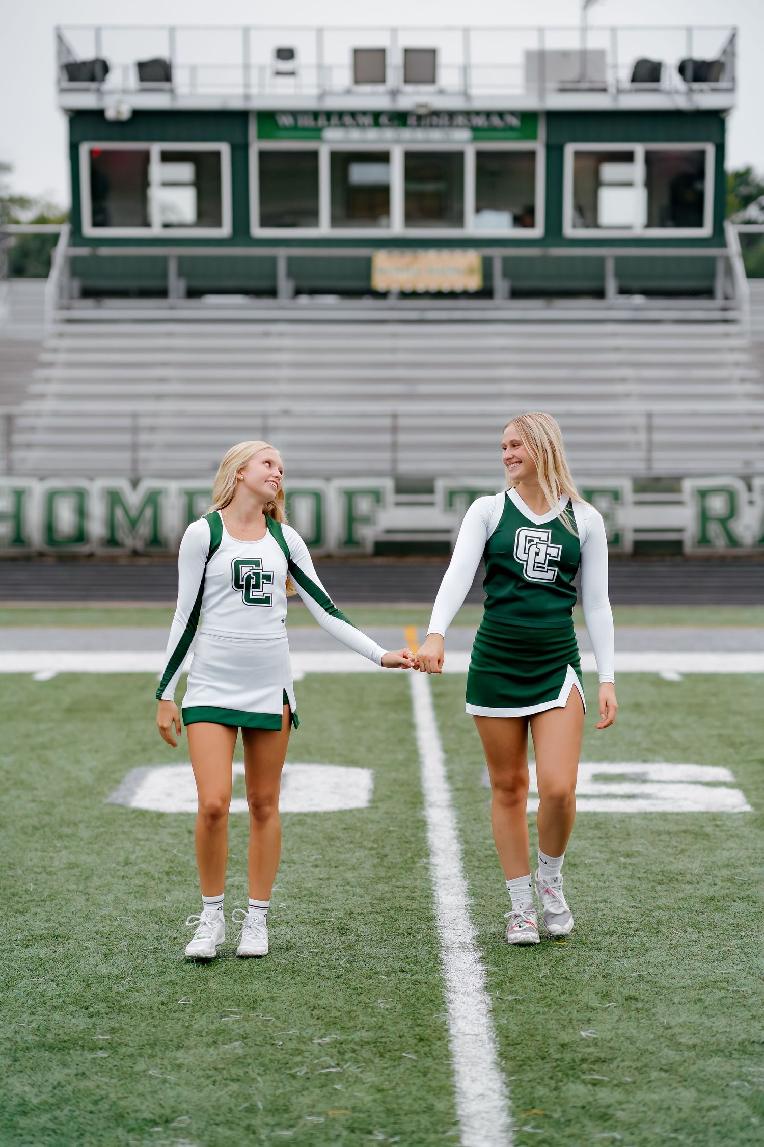 Two cheerleaders in green and white uniforms hold hands while walking on a football field in front of bleachers.