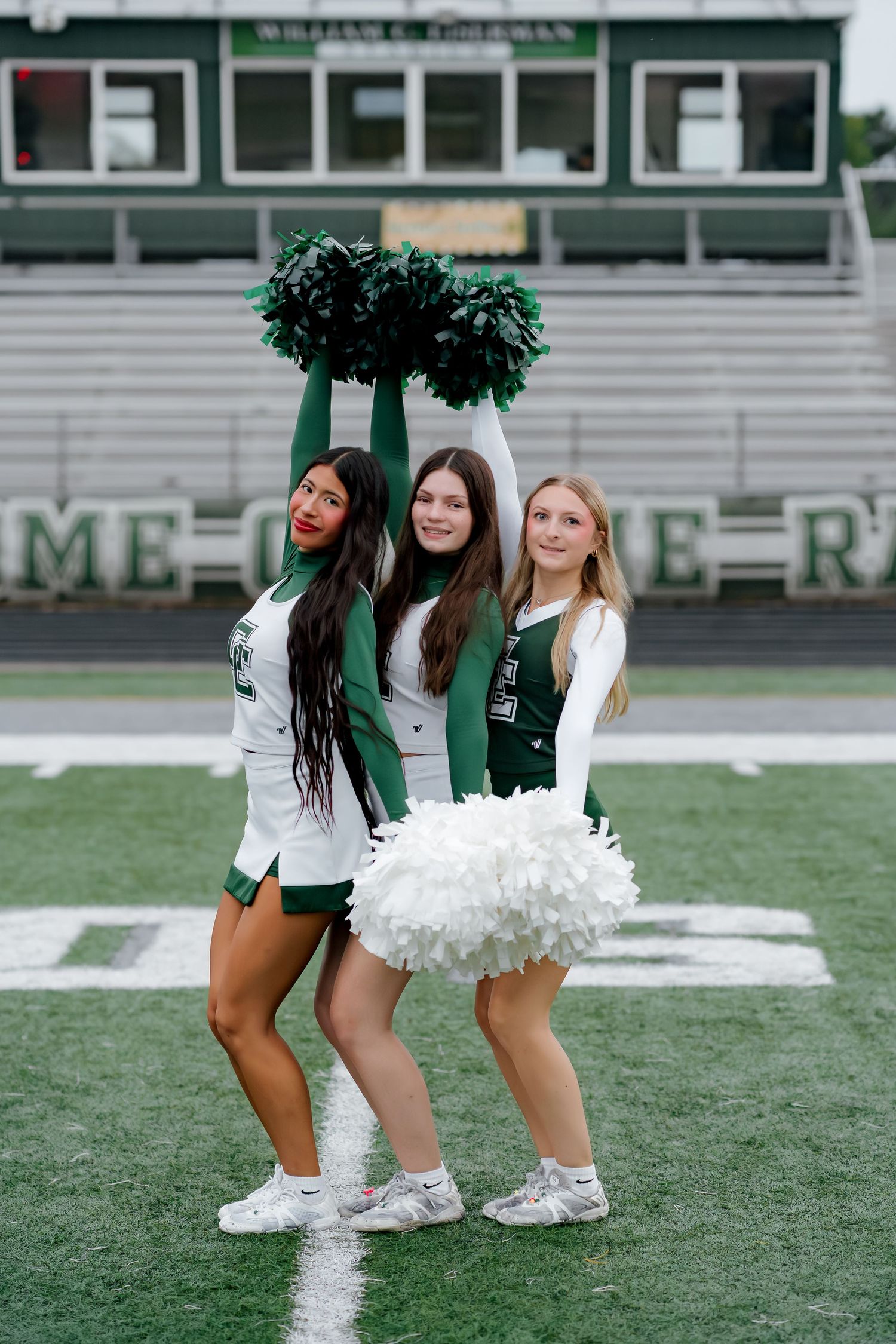 Three cheerleaders in green and white uniforms pose with pom-poms on a football field stadium.
