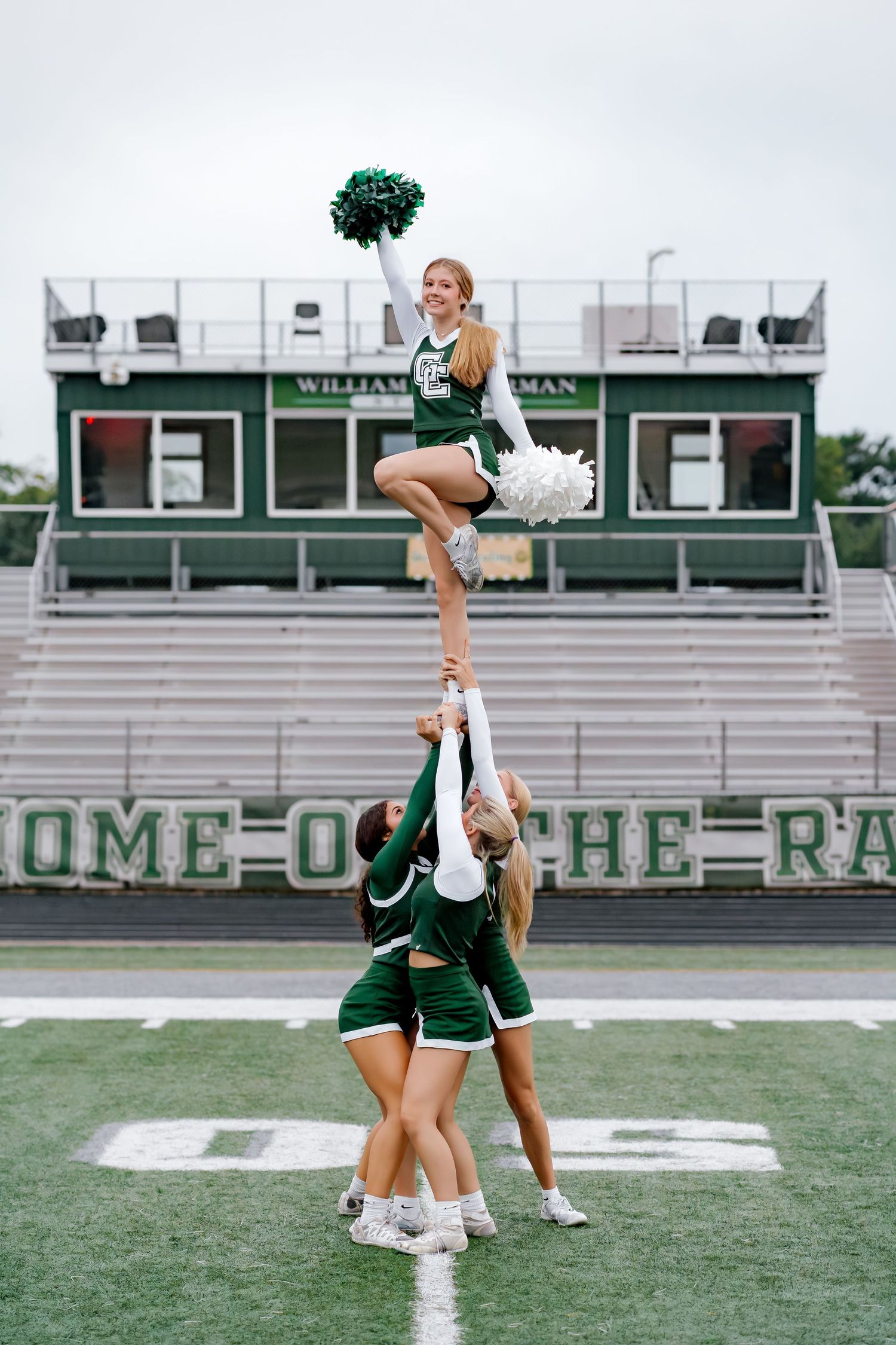 Cheerleaders perform a stunt at a football stadium, with one flyer lifted high holding green and white pom-poms.