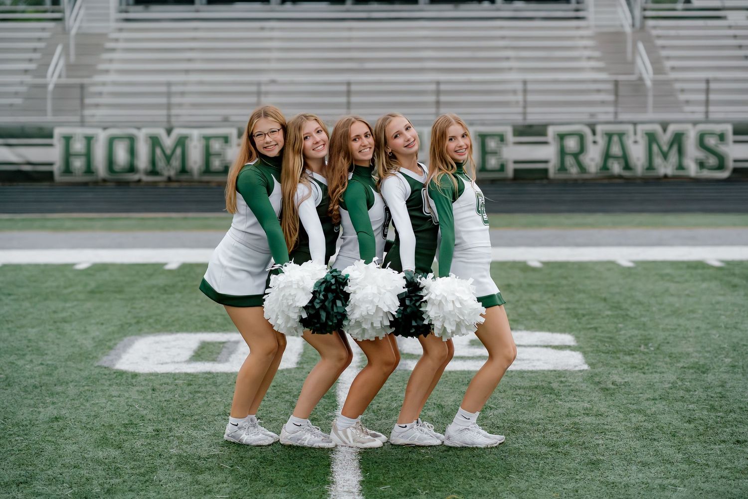 Five cheerleaders in green and white uniforms pose with pom-poms on a football field in front of a Rams sign.