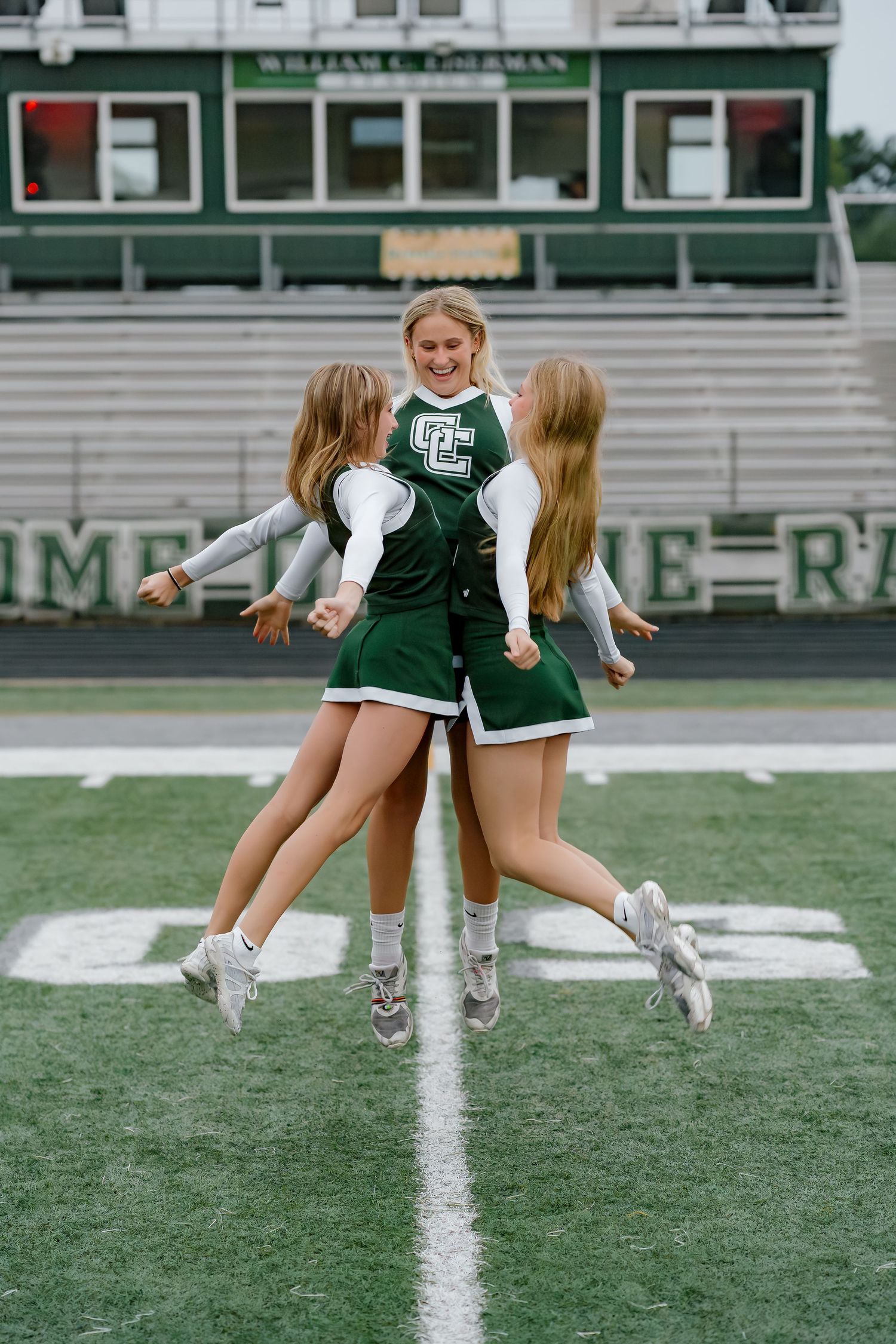 Three cheerleaders in green and white uniforms jumping together on a football field stadium.