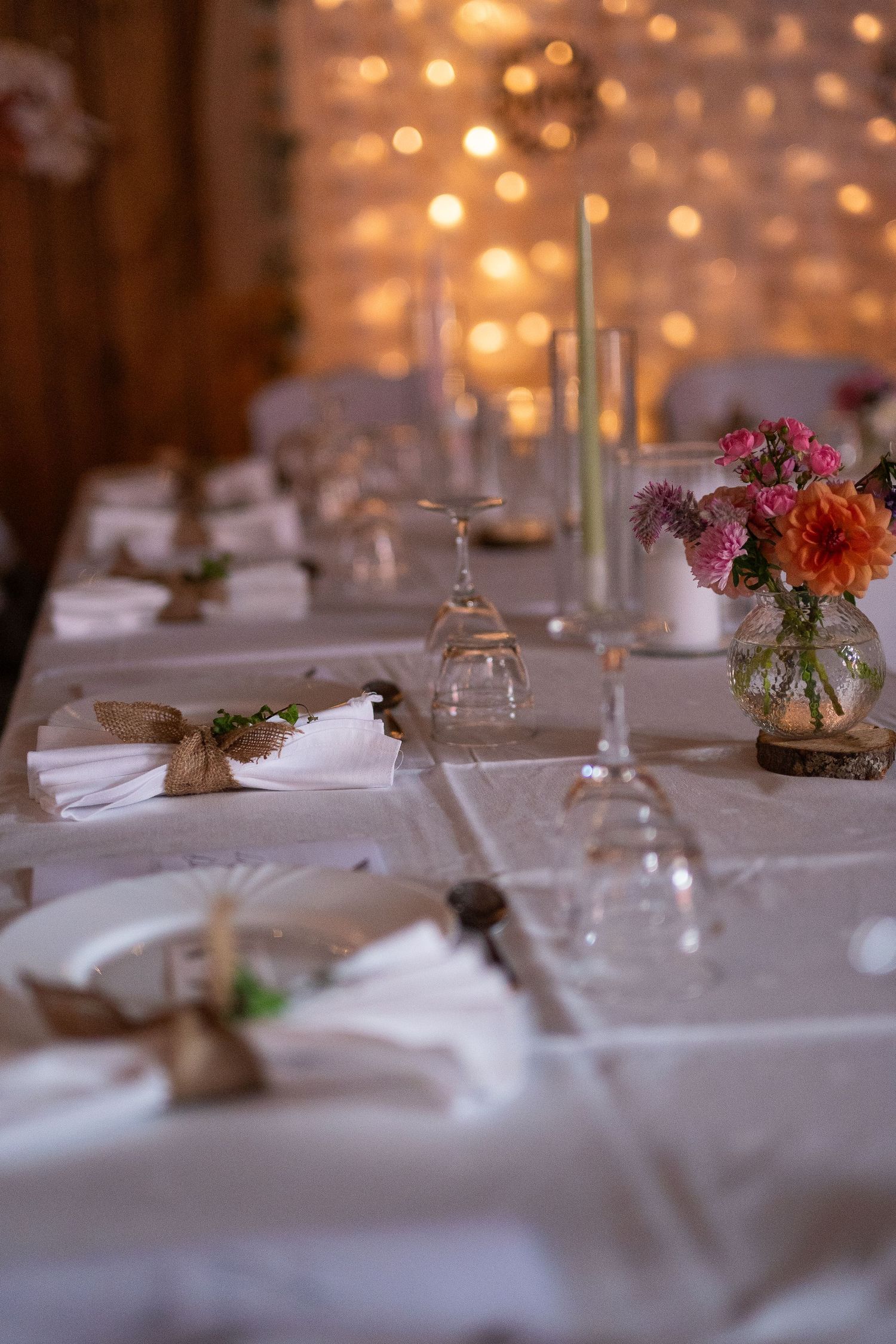 Bride smiling at candlelit wedding reception table with fairy lights and greenery backdrop in rustic barn venue.