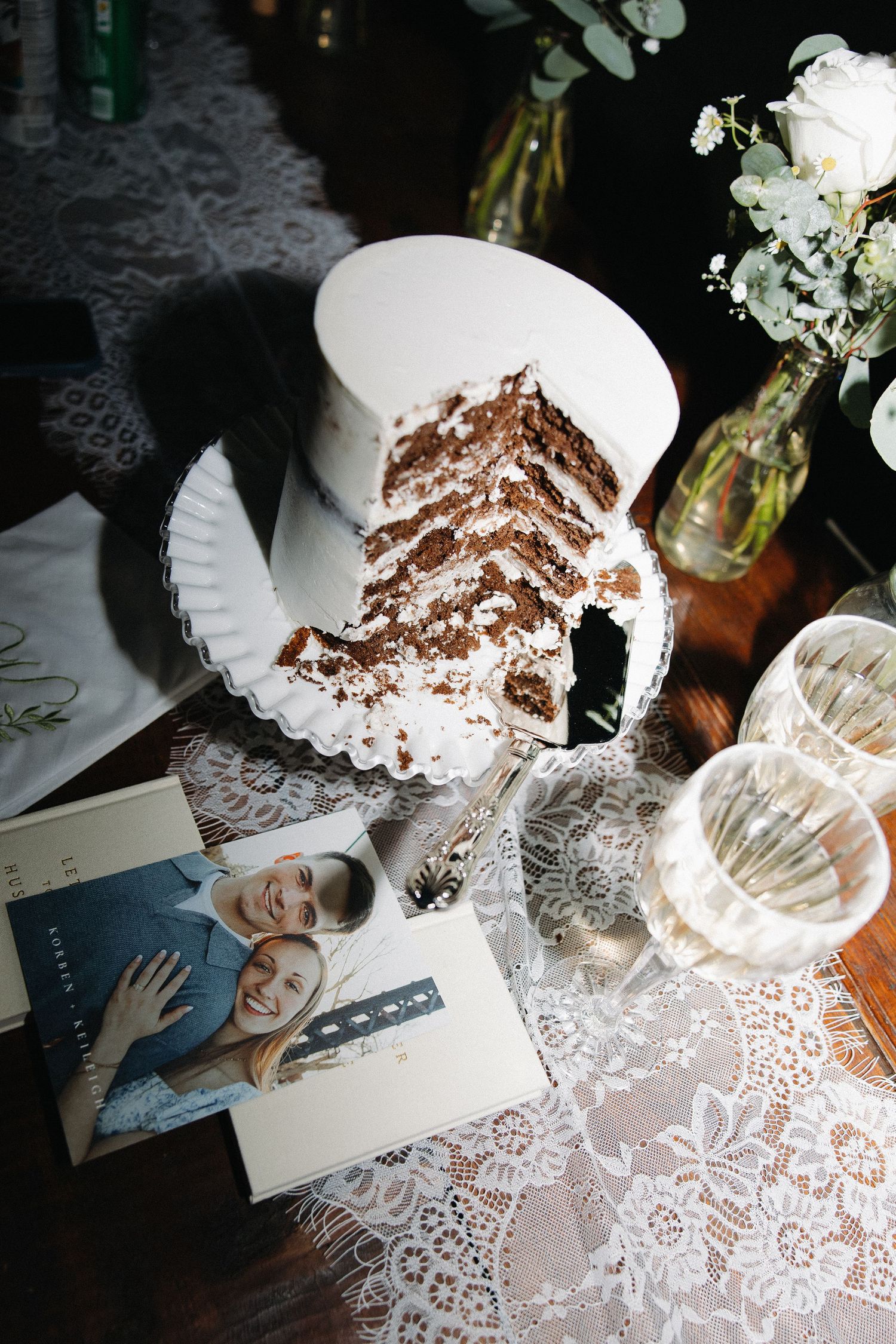Sliced white wedding cake on lace tablecloth with couple photo card and floral decor.