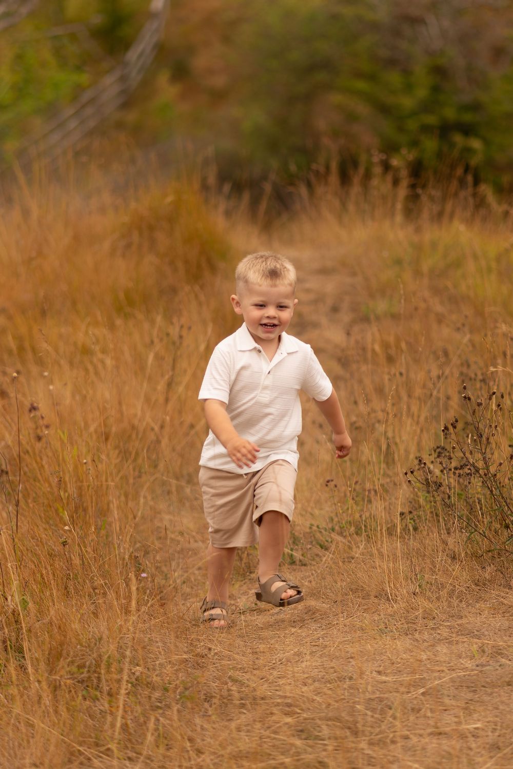 Young blonde boy running happily along a dirt path through golden autumn grass fields.