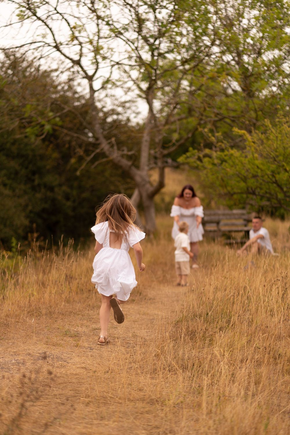 A young girl in a white dress runs toward her family on a sunny dirt path surrounded by tall grass and trees.