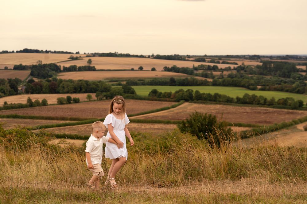 Two children in white outfits running through a grassy hillside with scenic English countryside fields in the background.
