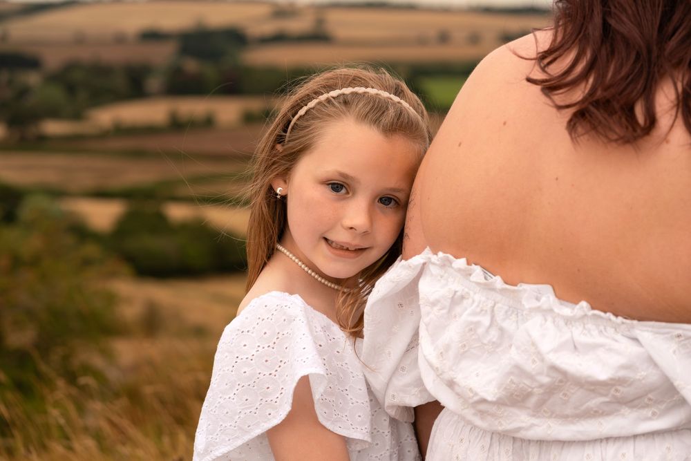 Young girl in white dress hugging pregnant mother's belly outdoors in scenic countryside setting.