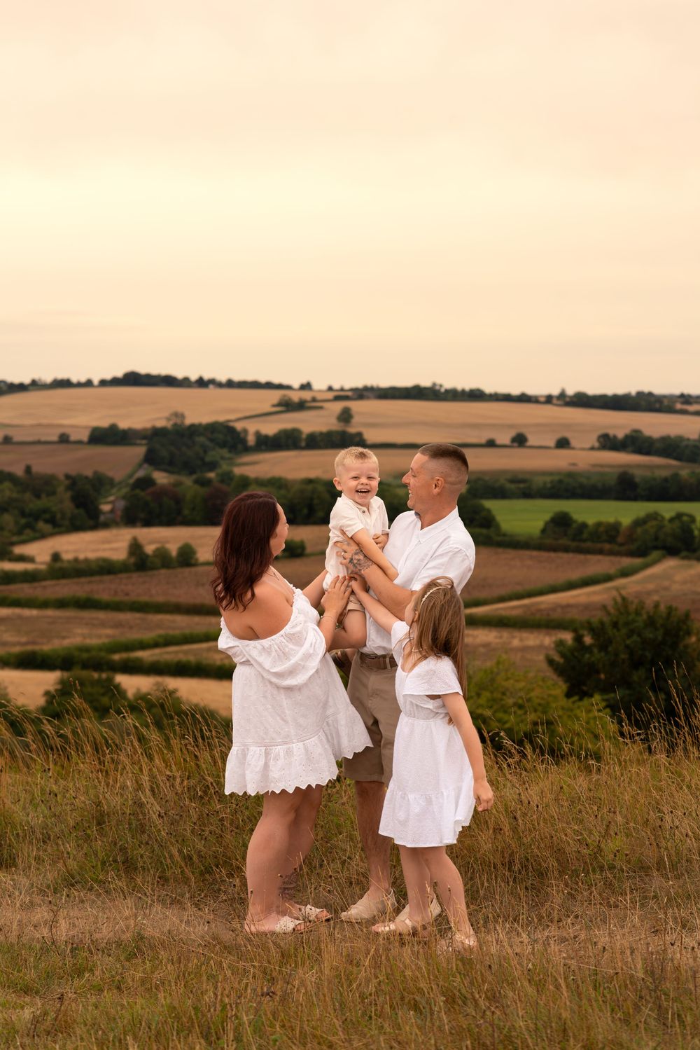 Family of four in white outfits posing on a hillside with scenic countryside fields at golden hour.