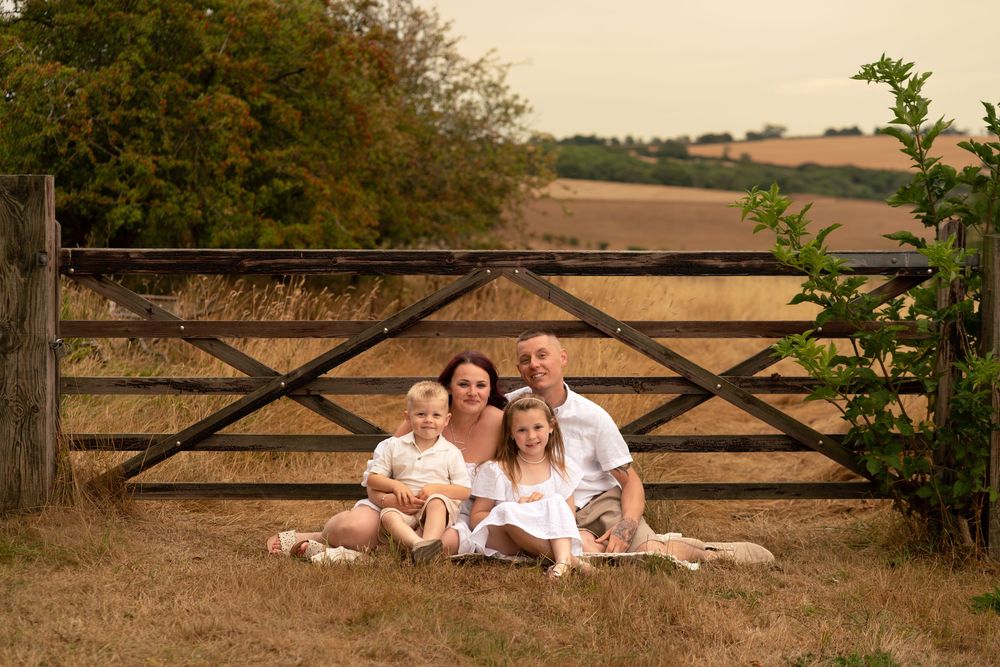 Family of four sitting in front of a wooden farm gate in a golden field, smiling together on a warm summer day.