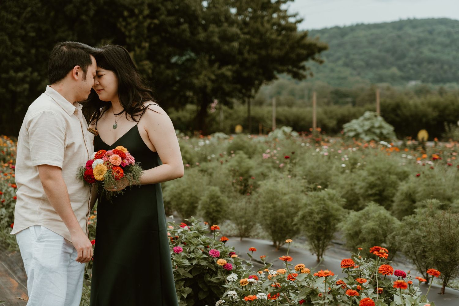 Couple embracing in a colorful flower garden with rolling green hills in the background, woman holding vibrant bouquet.
