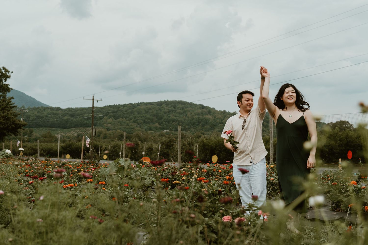 Couple walking joyfully through a colorful flower field with mountains in the background on a cloudy day.