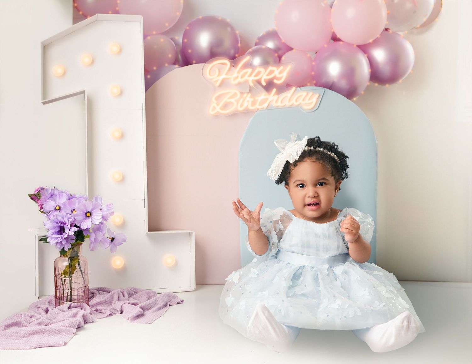 A baby girl in a white dress celebrates her first birthday with pink balloons and a Happy Birthday neon sign.