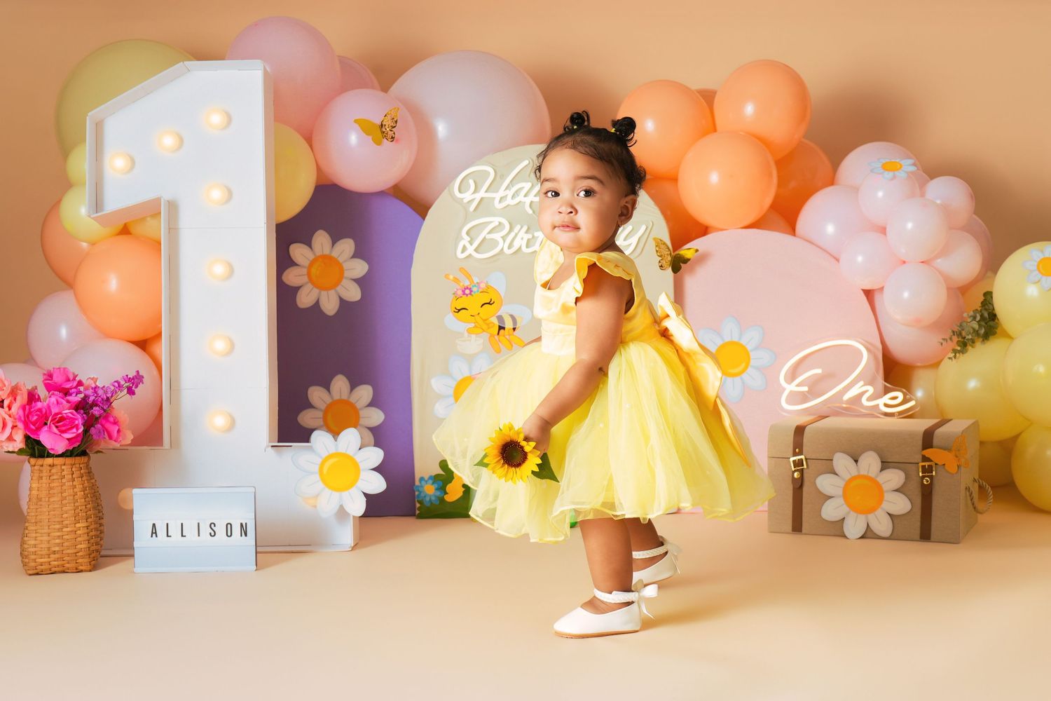 Toddler girl in yellow dress at first birthday party with balloon arch, daisy decorations, and number one backdrop.