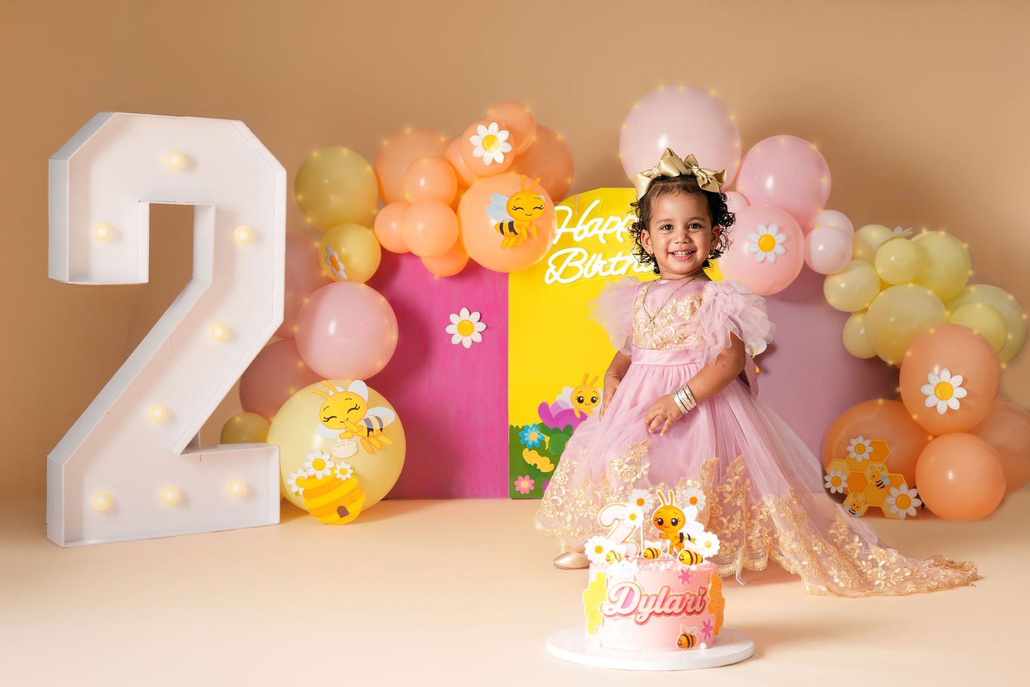 Toddler girl in pink dress celebrates 2nd birthday with balloon arch, floral cake, and lit number 2 decoration.