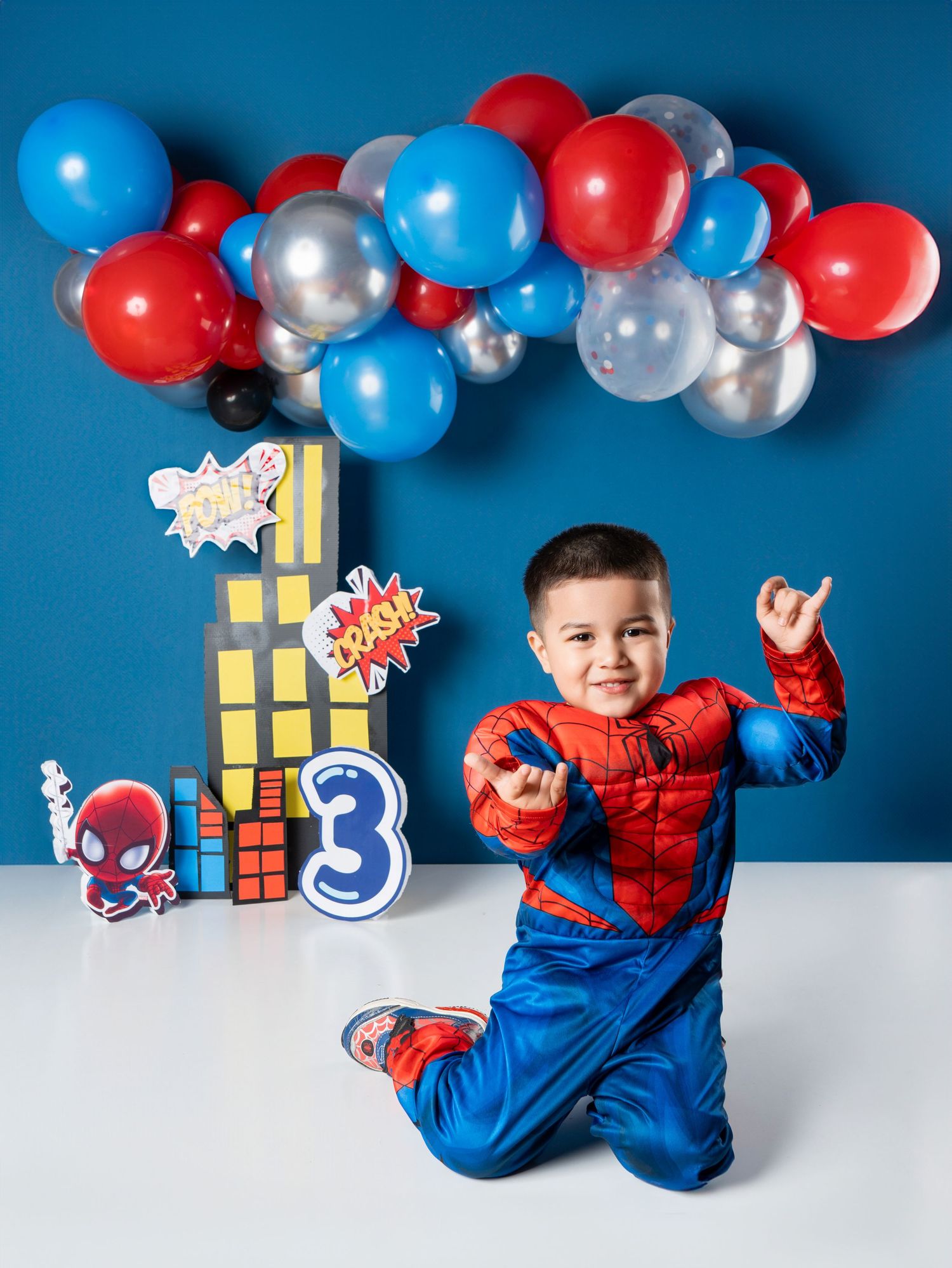 Toddler in Spiderman costume poses at 3rd birthday party with red, blue, silver balloons and comic-themed decorations.