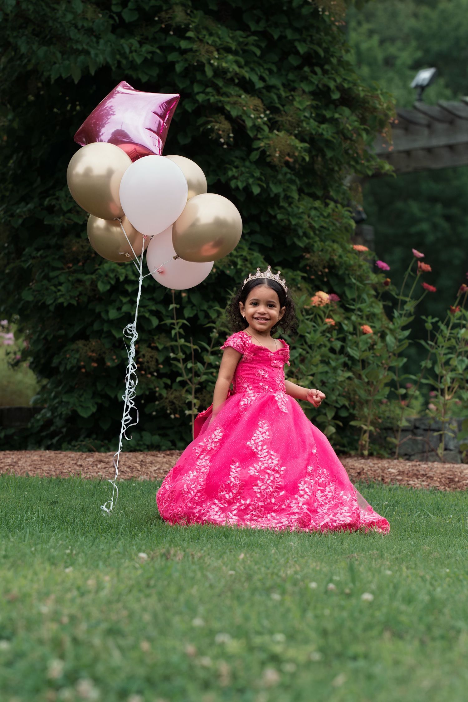 Little girl in pink princess gown with tiara holds colorful balloons outdoors on green lawn.