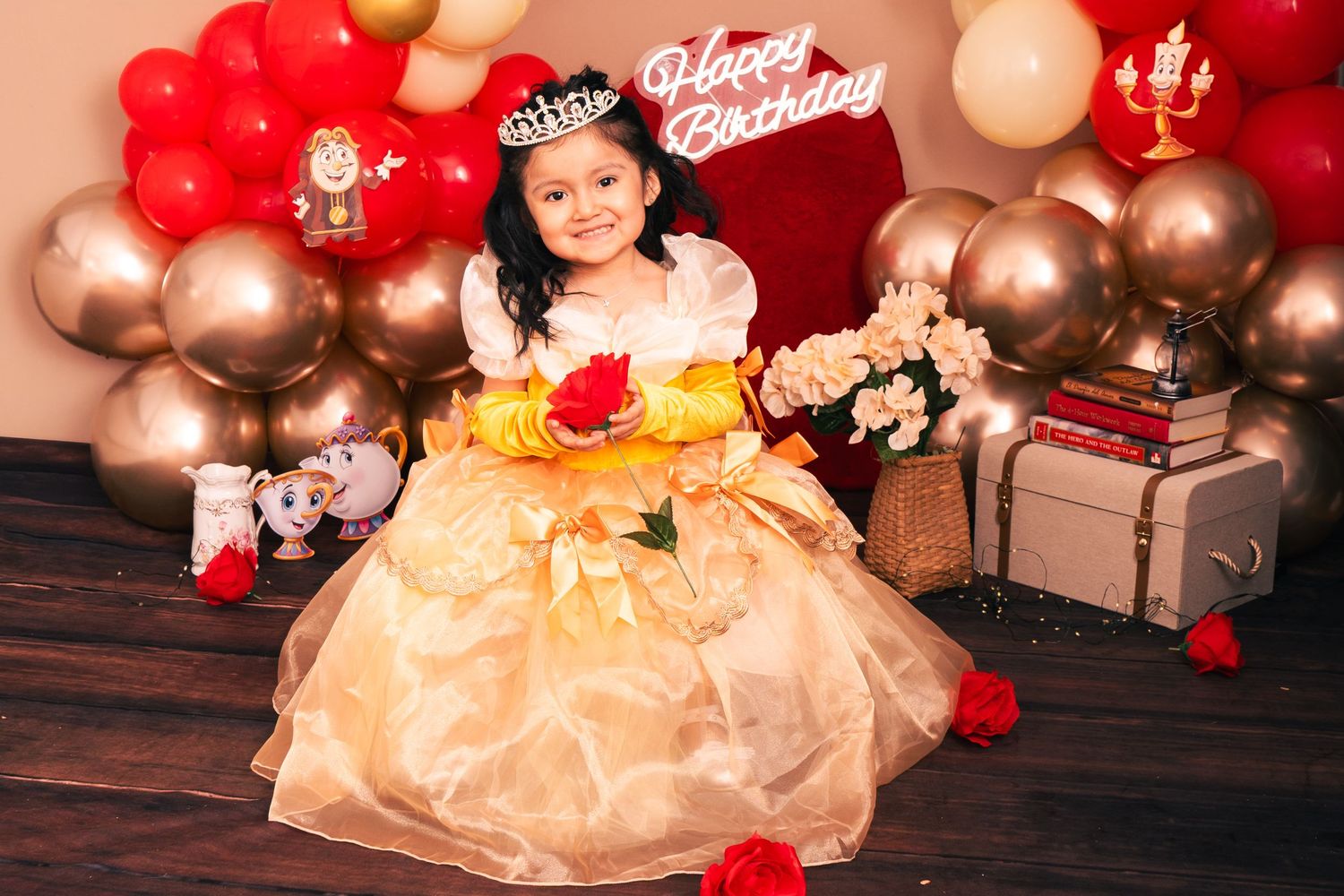 A little girl in a golden Belle princess dress poses at a Beauty and the Beast themed birthday party with balloons.