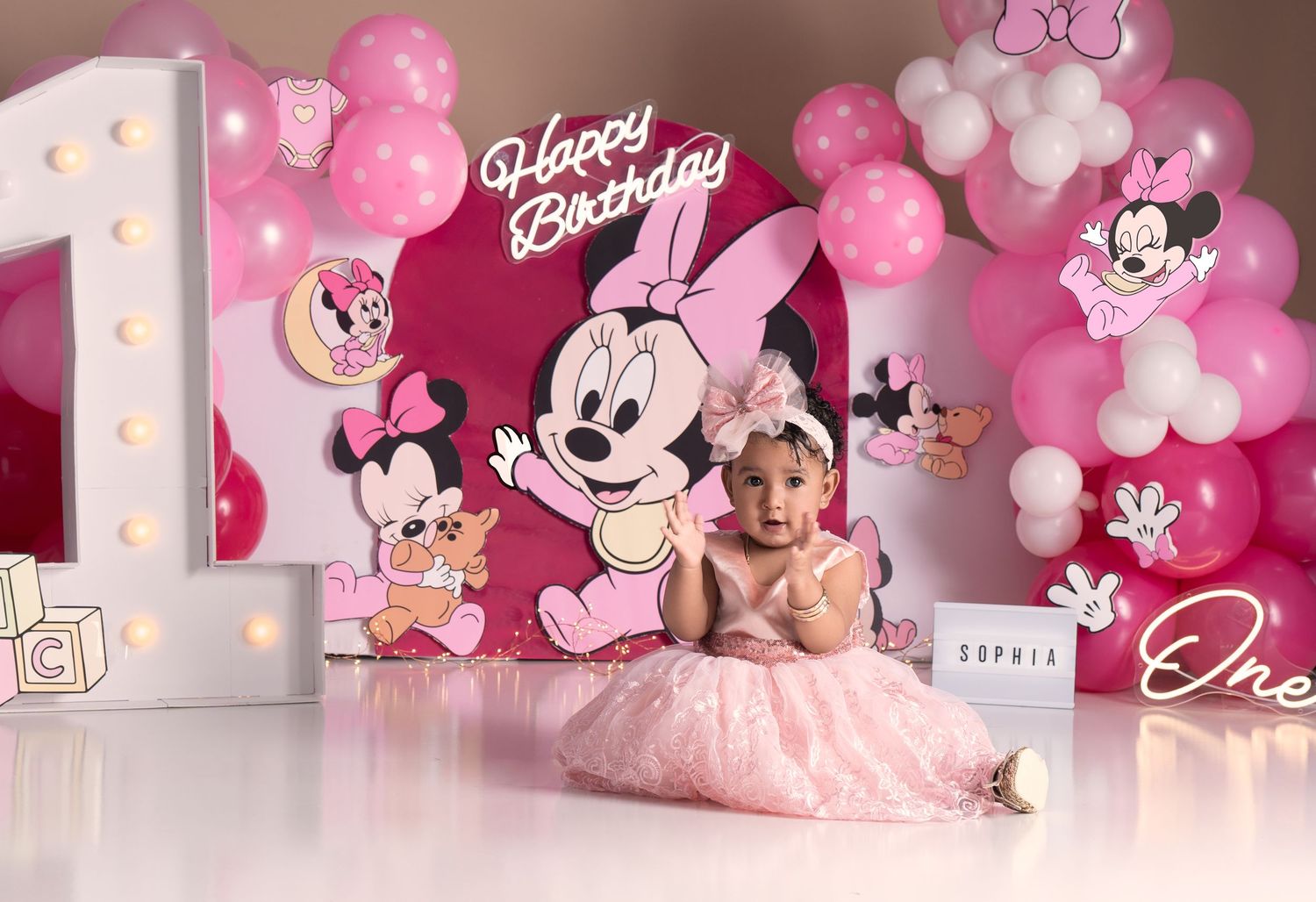 Baby girl in pink tutu sits before Minnie Mouse Happy Birthday backdrop with pink balloons for 1st birthday.