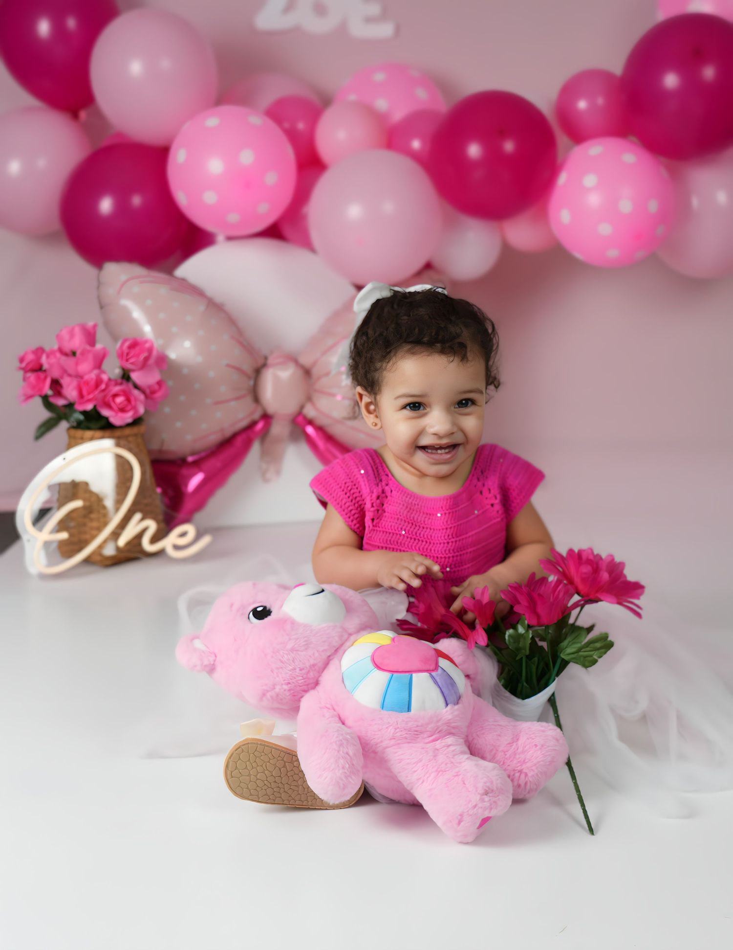 Baby girl in pink dress smiles at first birthday party with balloons, Care Bear toy, and roses.