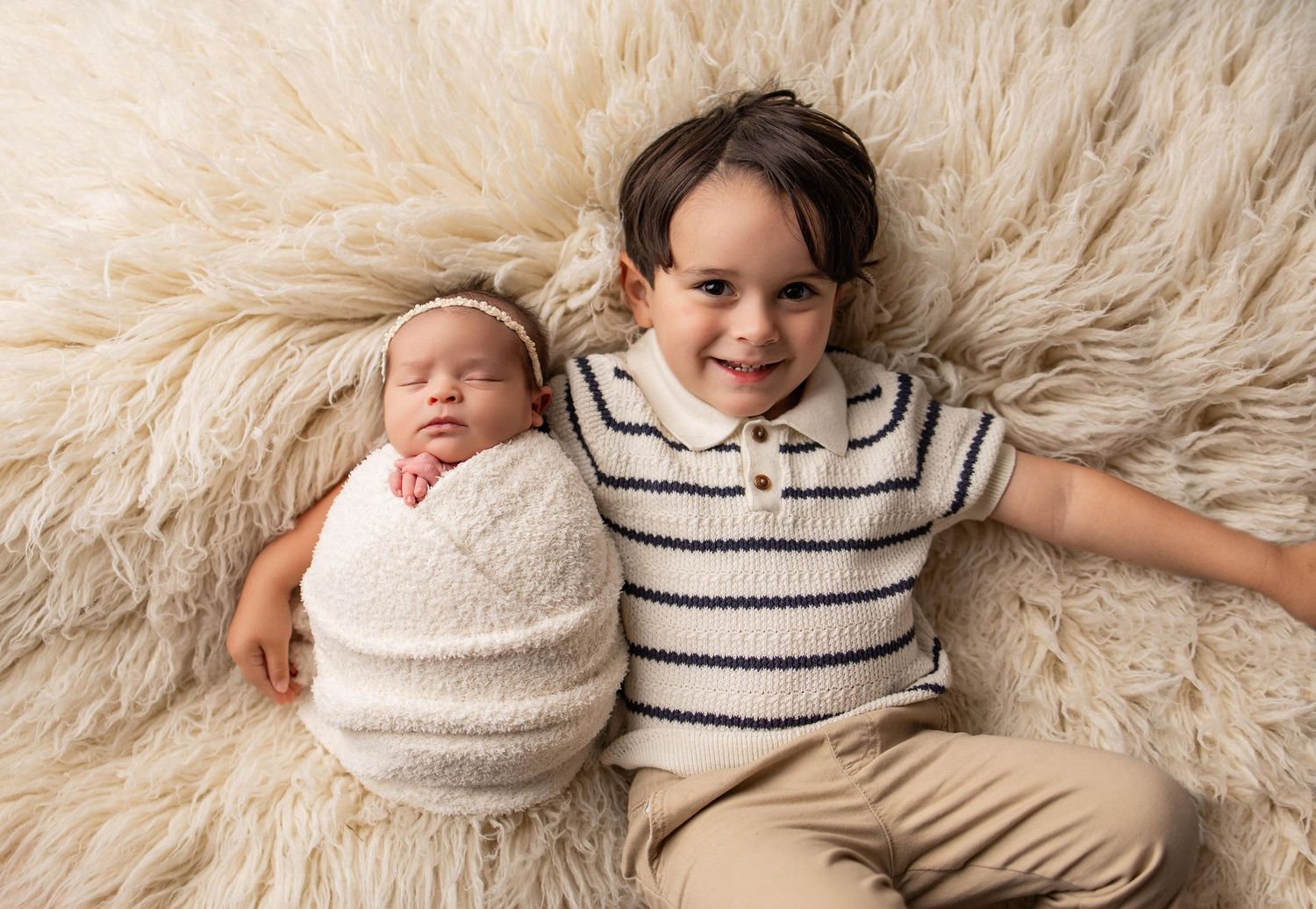 A smiling toddler boy in a striped shirt poses with a sleeping newborn baby wrapped in white on a fluffy cream rug.