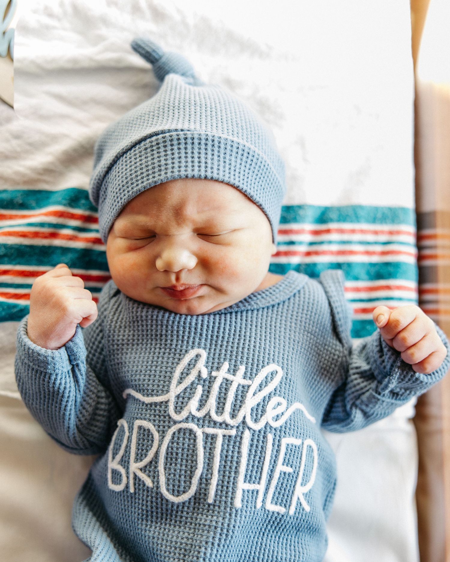 Newborn baby wearing a blue waffle knit little brother outfit and matching knotted hat, lying on a striped blanket.