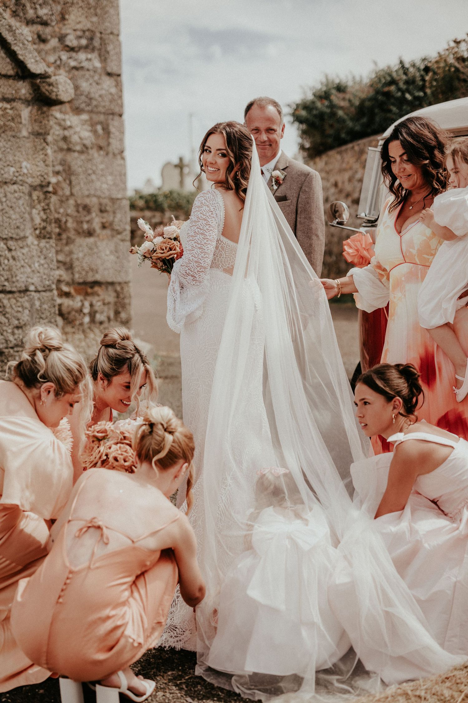 Bride in white gown and veil surrounded by bridesmaids in blush pink dresses outside a stone church.