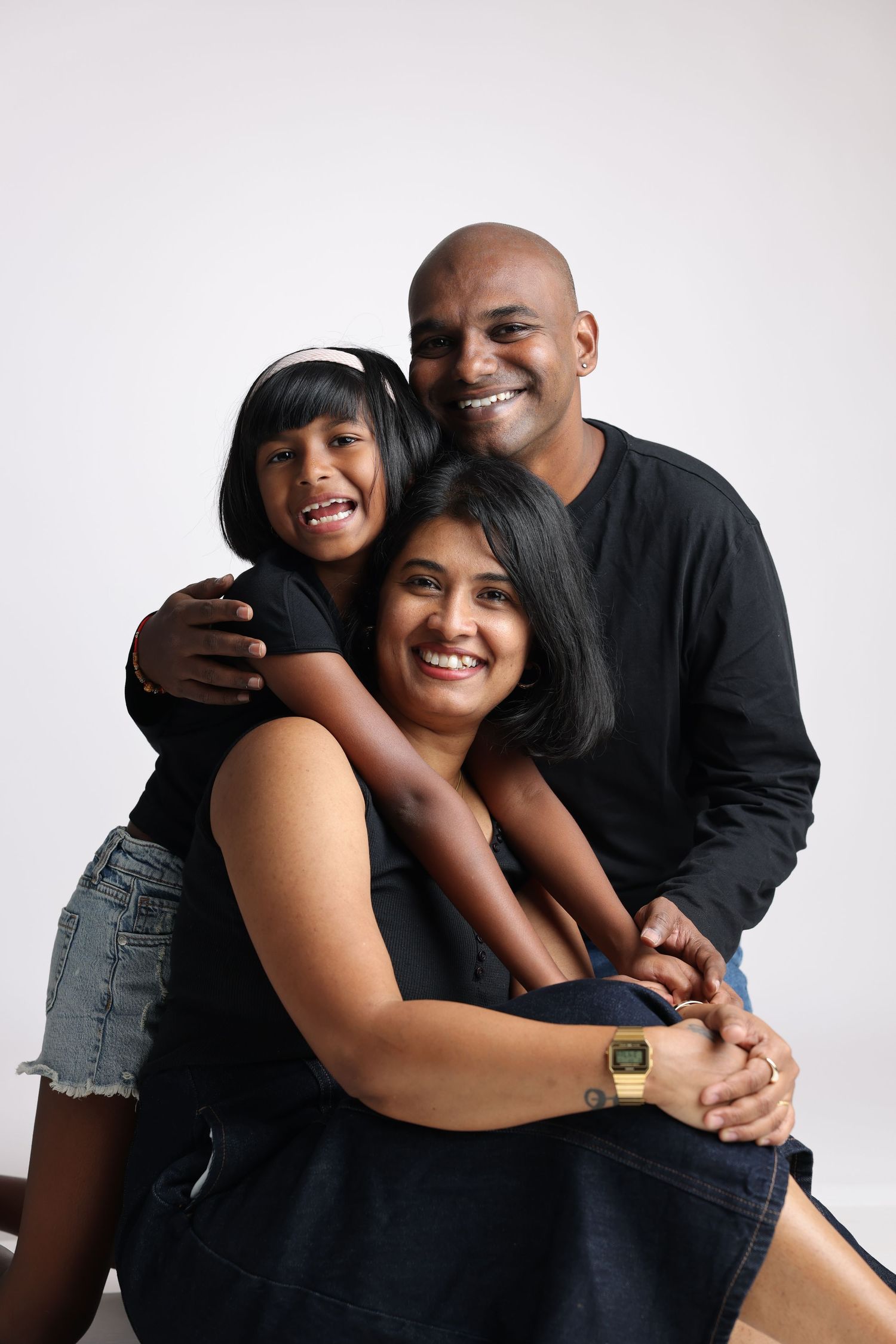 A smiling South Asian family of three in black outfits poses together, with a young girl hugging her parents warmly.
