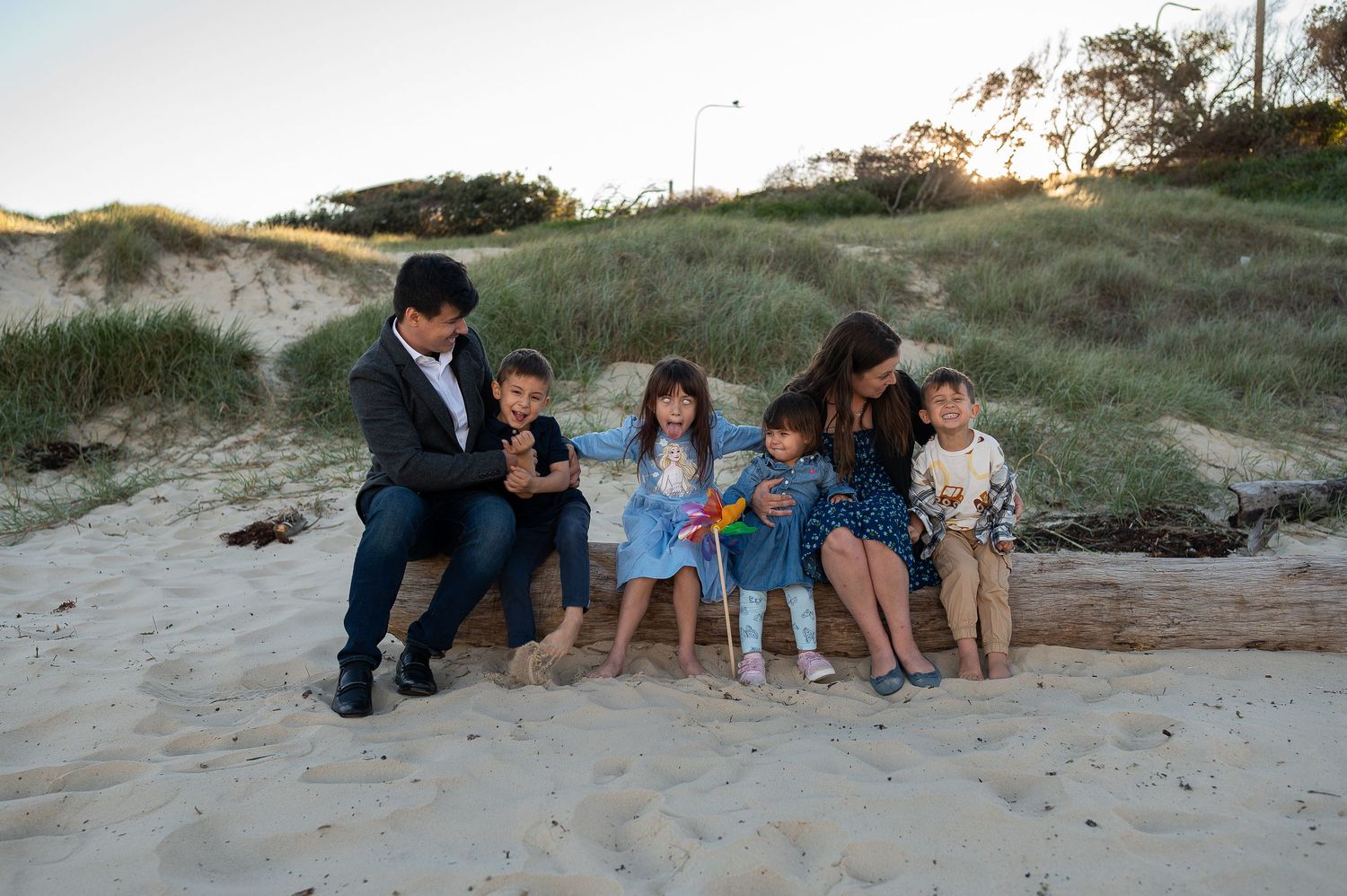Family of six sitting on driftwood on a sandy beach with grass-covered dunes in the background.