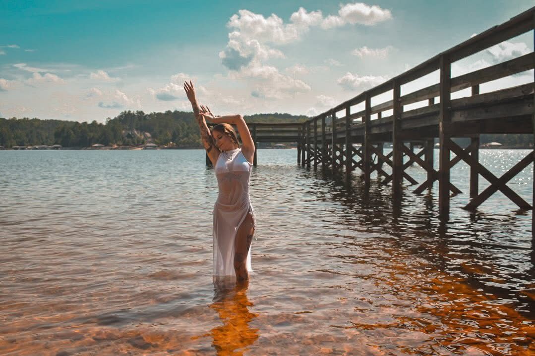 Woman in white swimsuit standing in lake water with arms raised near wooden pier on sunny day.