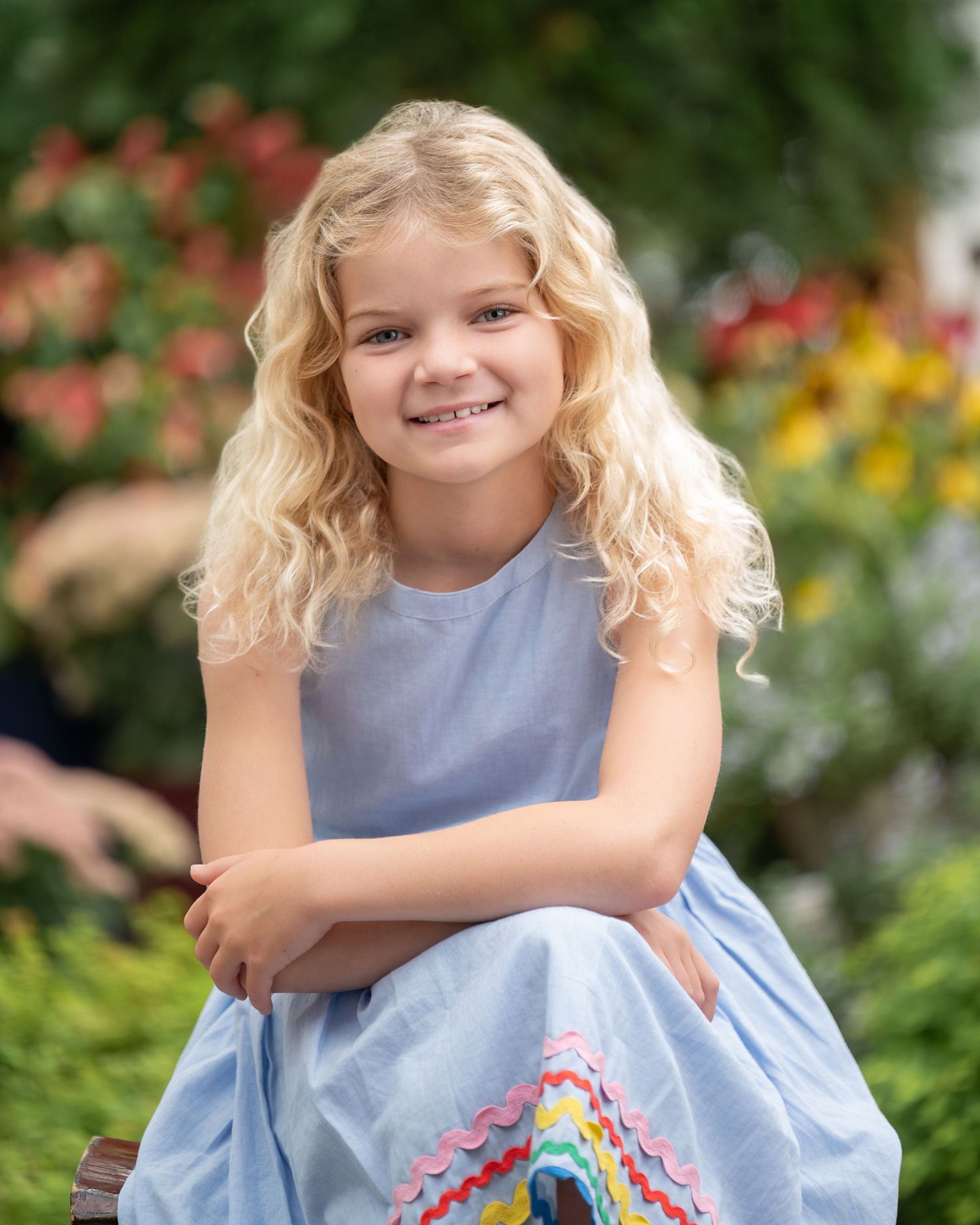 Young girl with curly blonde hair smiling in a blue dress, seated outdoors among colorful garden flowers.