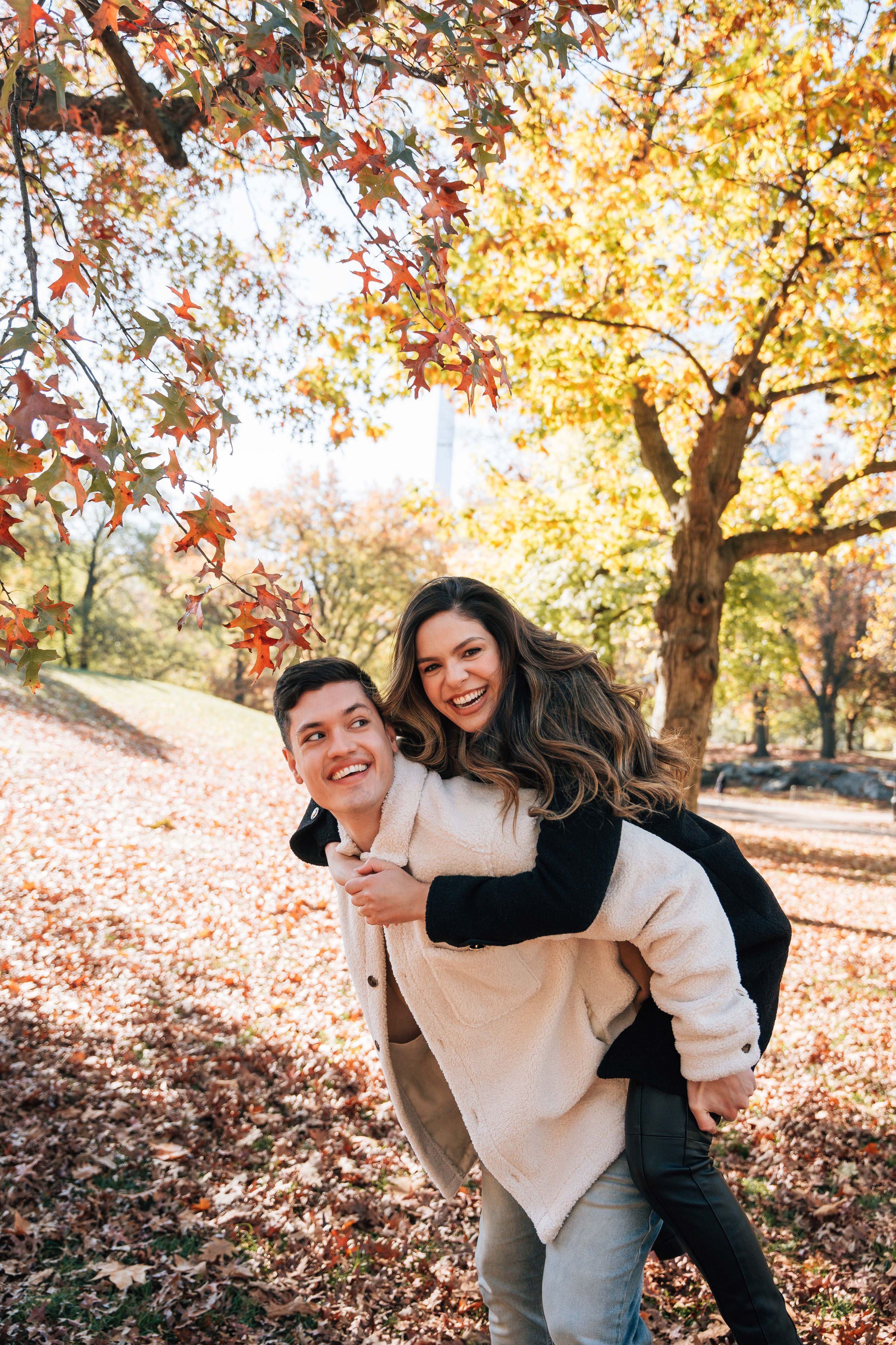 A happy couple enjoys a piggyback ride in an autumn park, surrounded by vibrant red and yellow fall foliage.