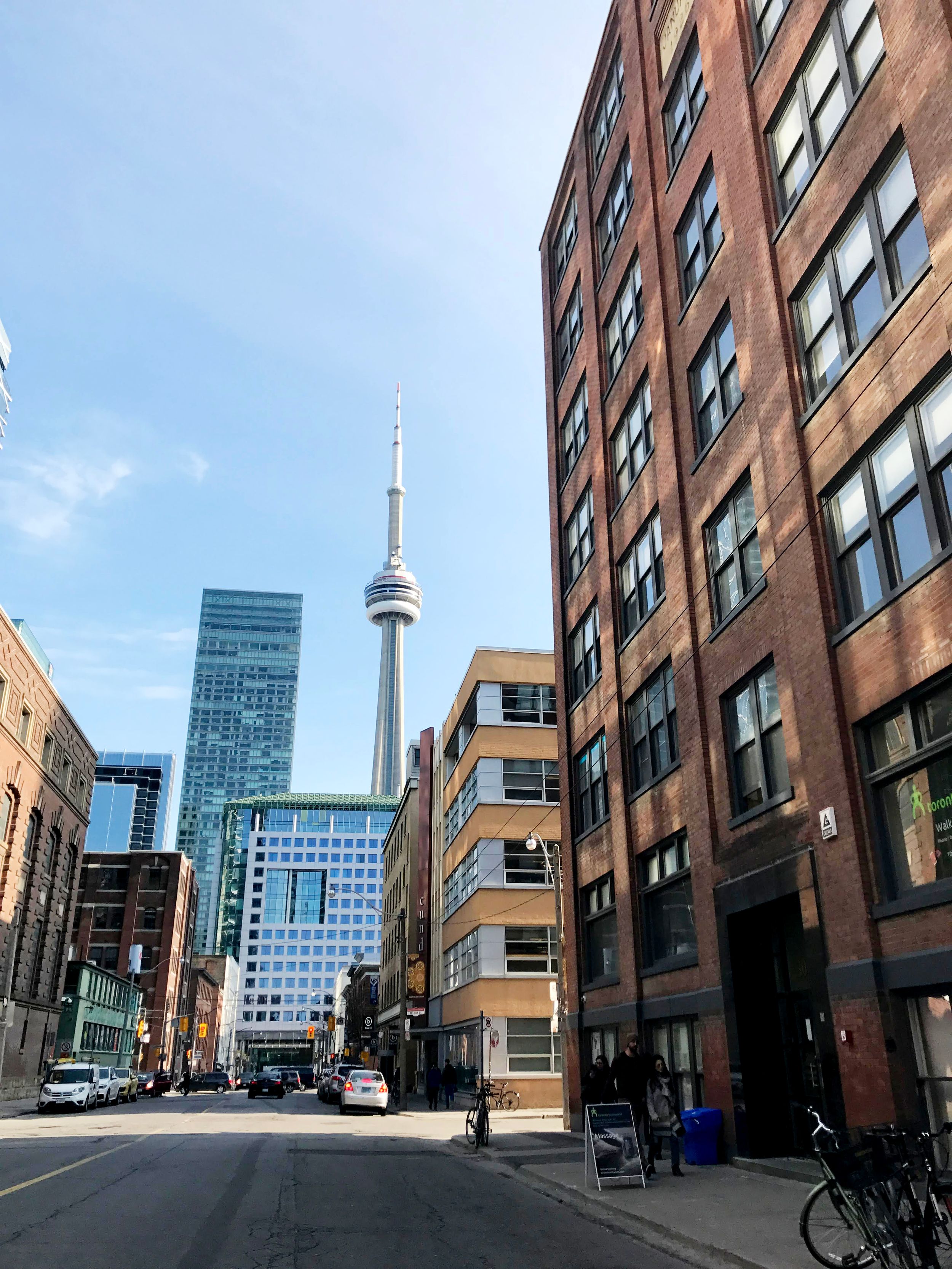 Street view of Toronto with CN Tower visible between brick buildings under a clear blue sky.