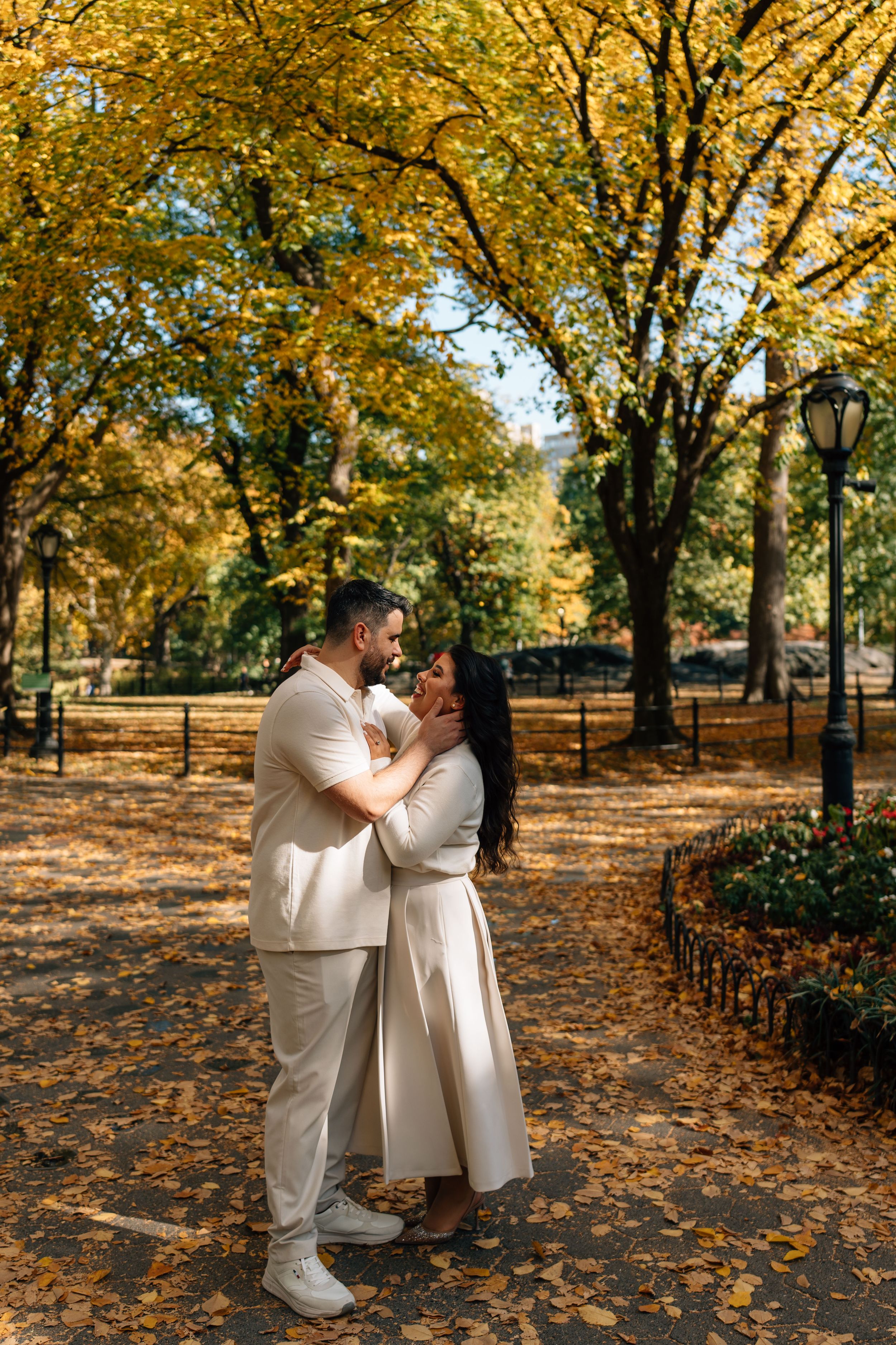 Couple embracing in autumn park with golden foliage, wearing matching cream outfits on a leaf-covered path.