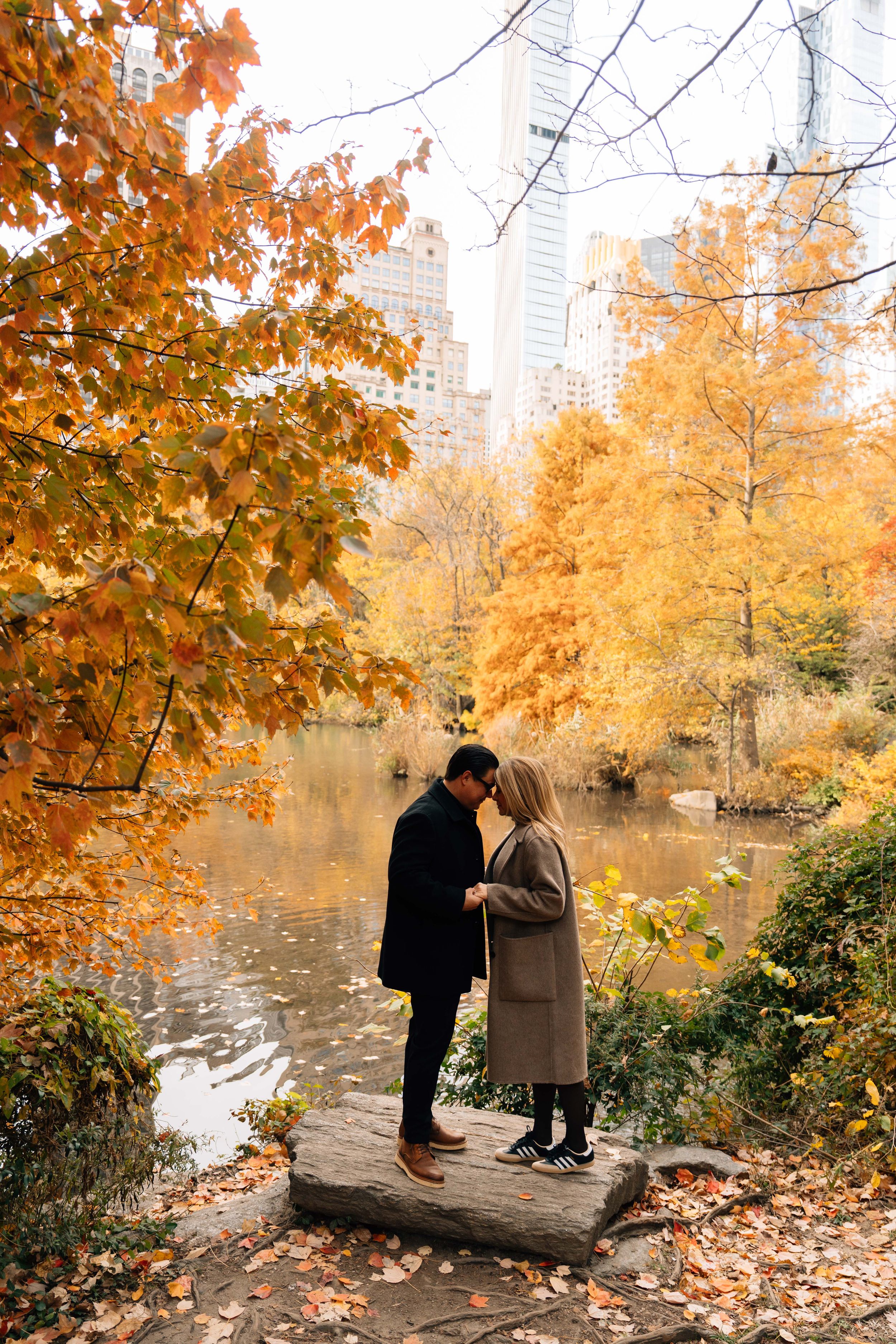 Couple sharing a romantic moment by a pond in Central Park surrounded by vibrant autumn foliage and NYC skyline.