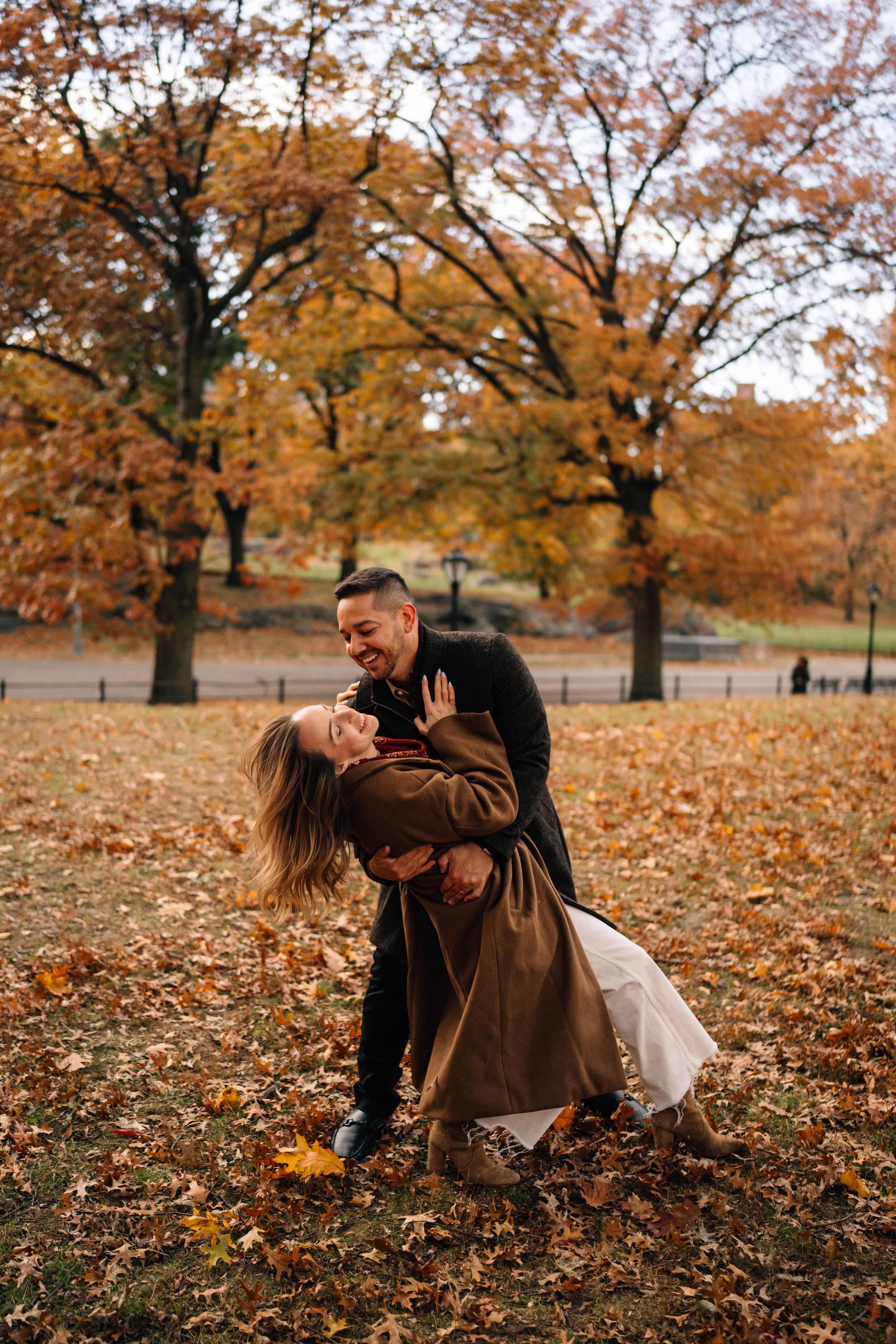 Couple sharing a romantic dip kiss in Central Park surrounded by vibrant autumn foliage and fallen leaves.