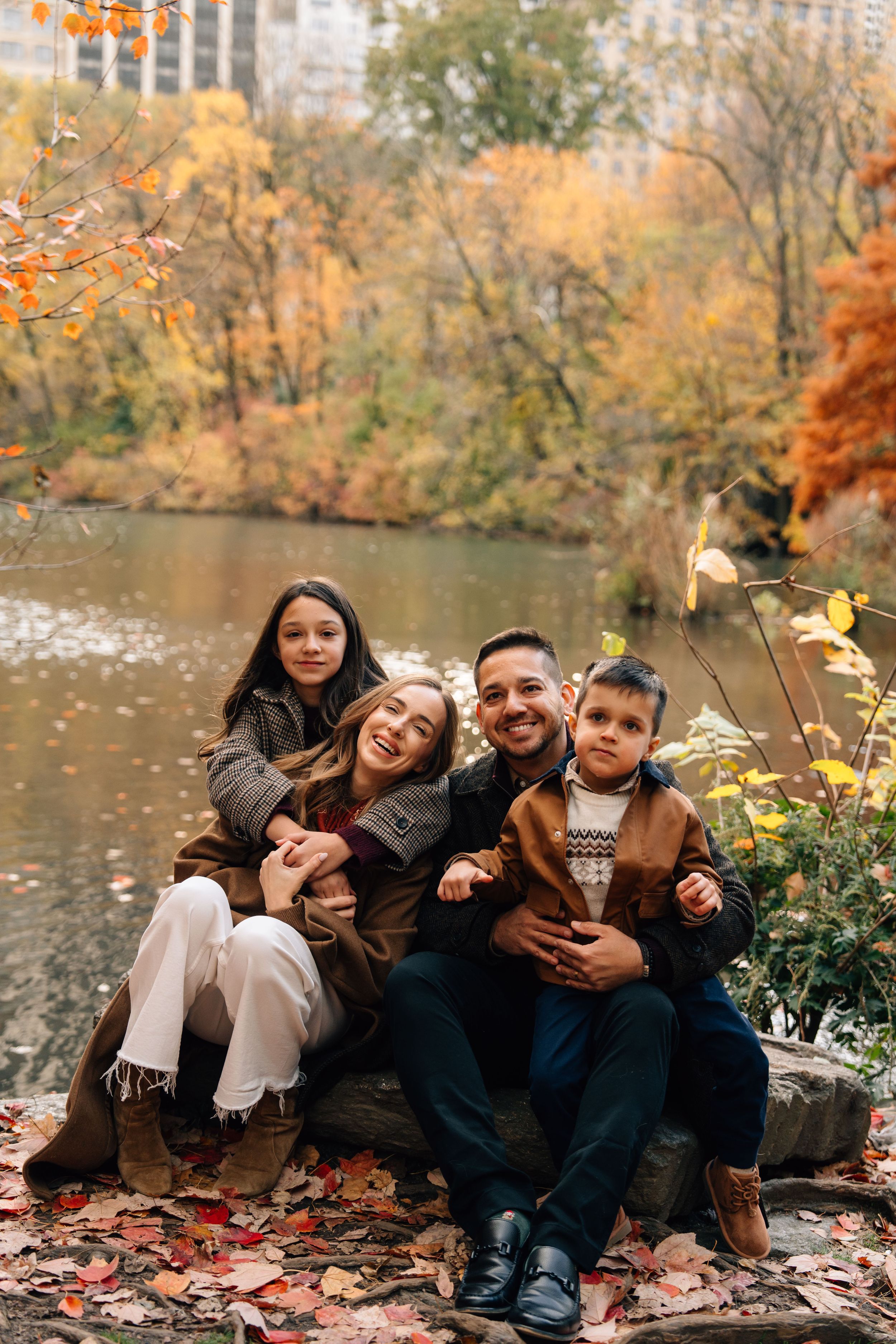 Happy family of four sitting by a pond in an autumn park, surrounded by colorful fall foliage.