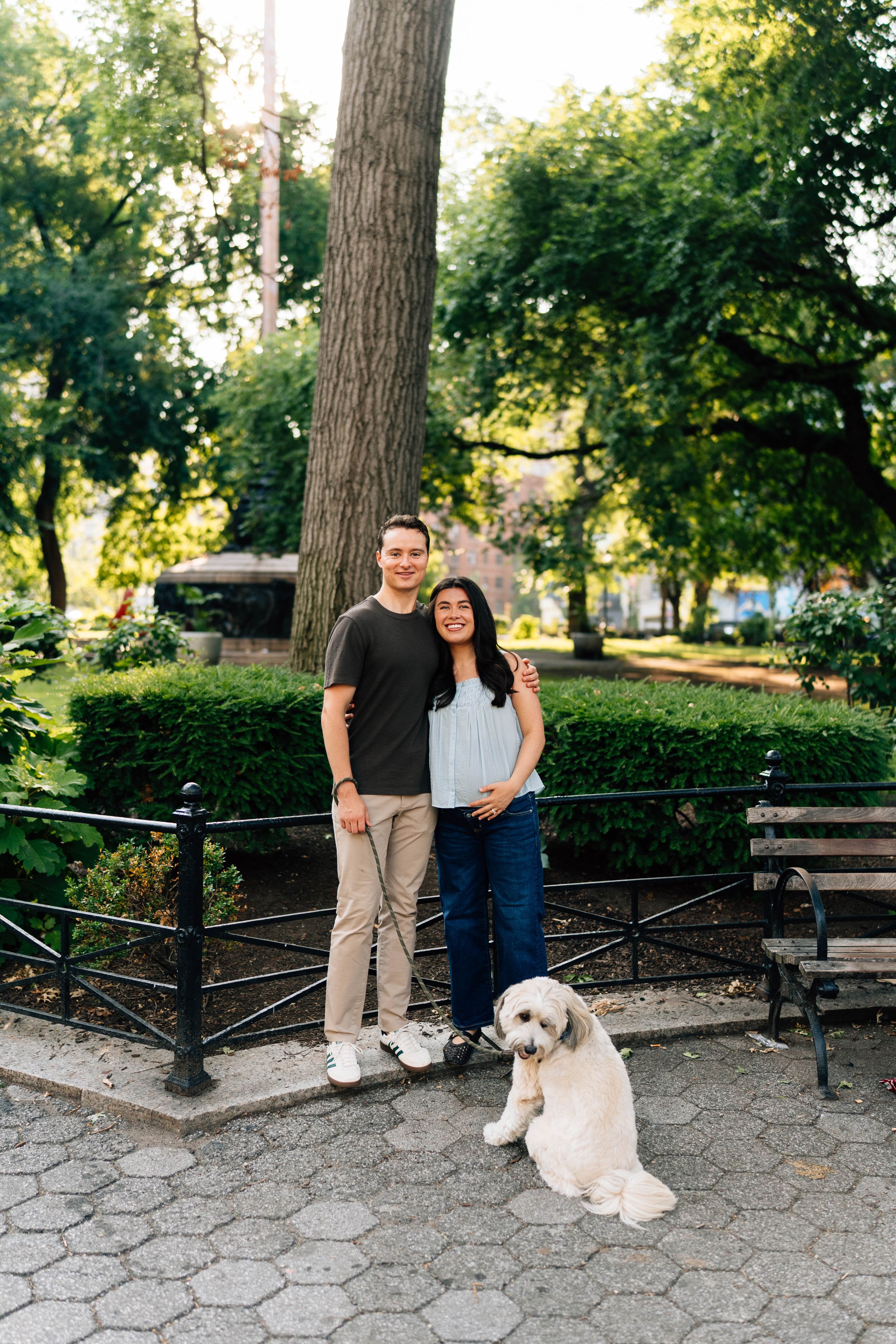 A couple smiles with their white fluffy dog in a lush green park, standing near a black iron fence and bench.